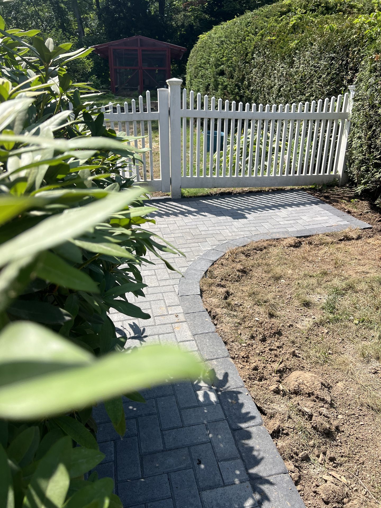 Curved brick walkway leading to a white picket gate in a garden, with green plants and a dry lawn.
