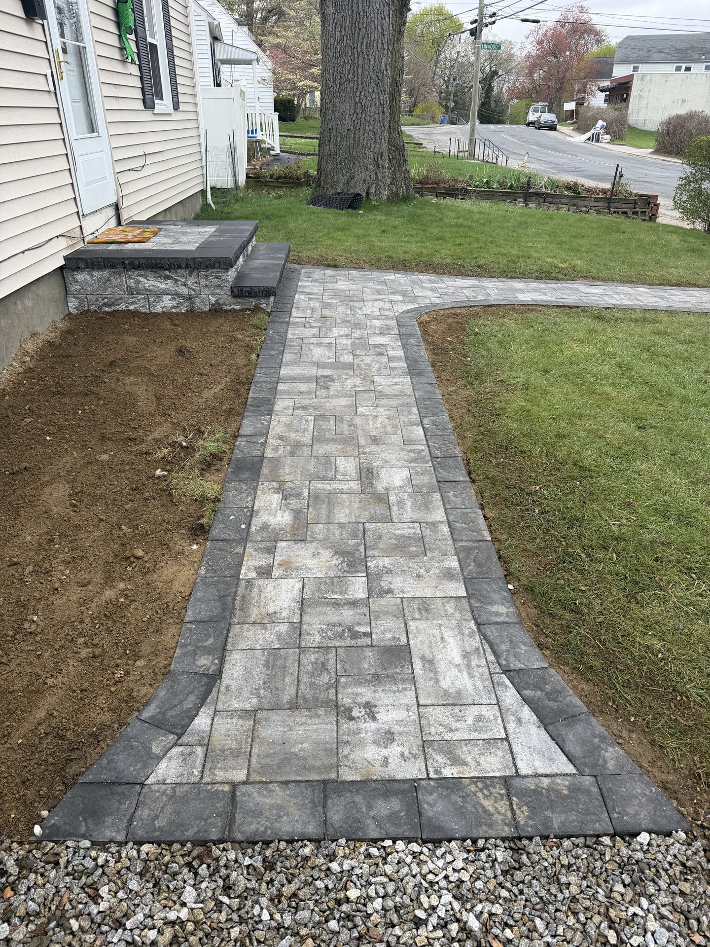 New gray paver walkway beside a house, bordered by grass and soil, leading to a front step