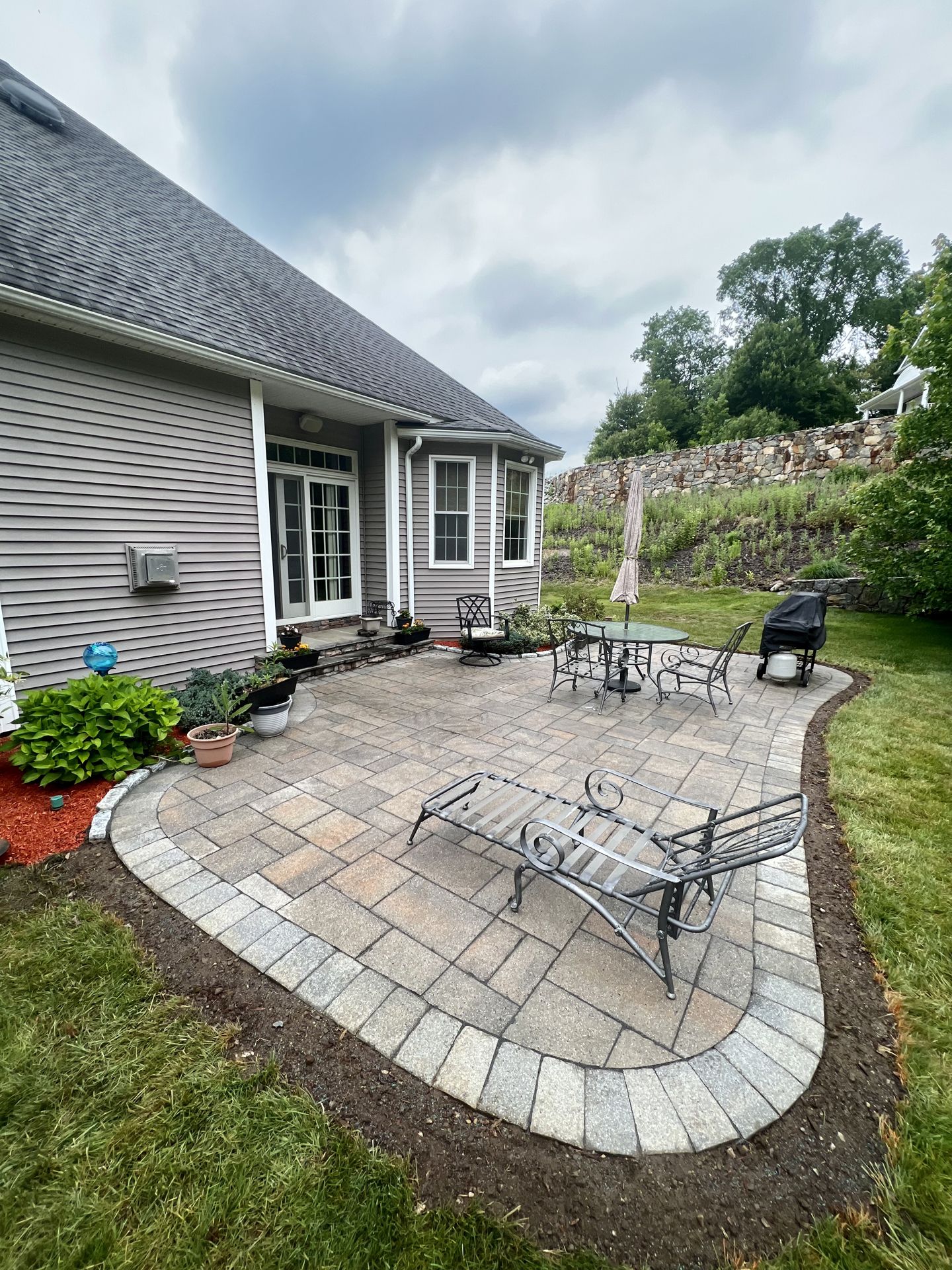 Back patio with stone pavers, metal chairs, barbecue grill, and house exterior under cloudy sky