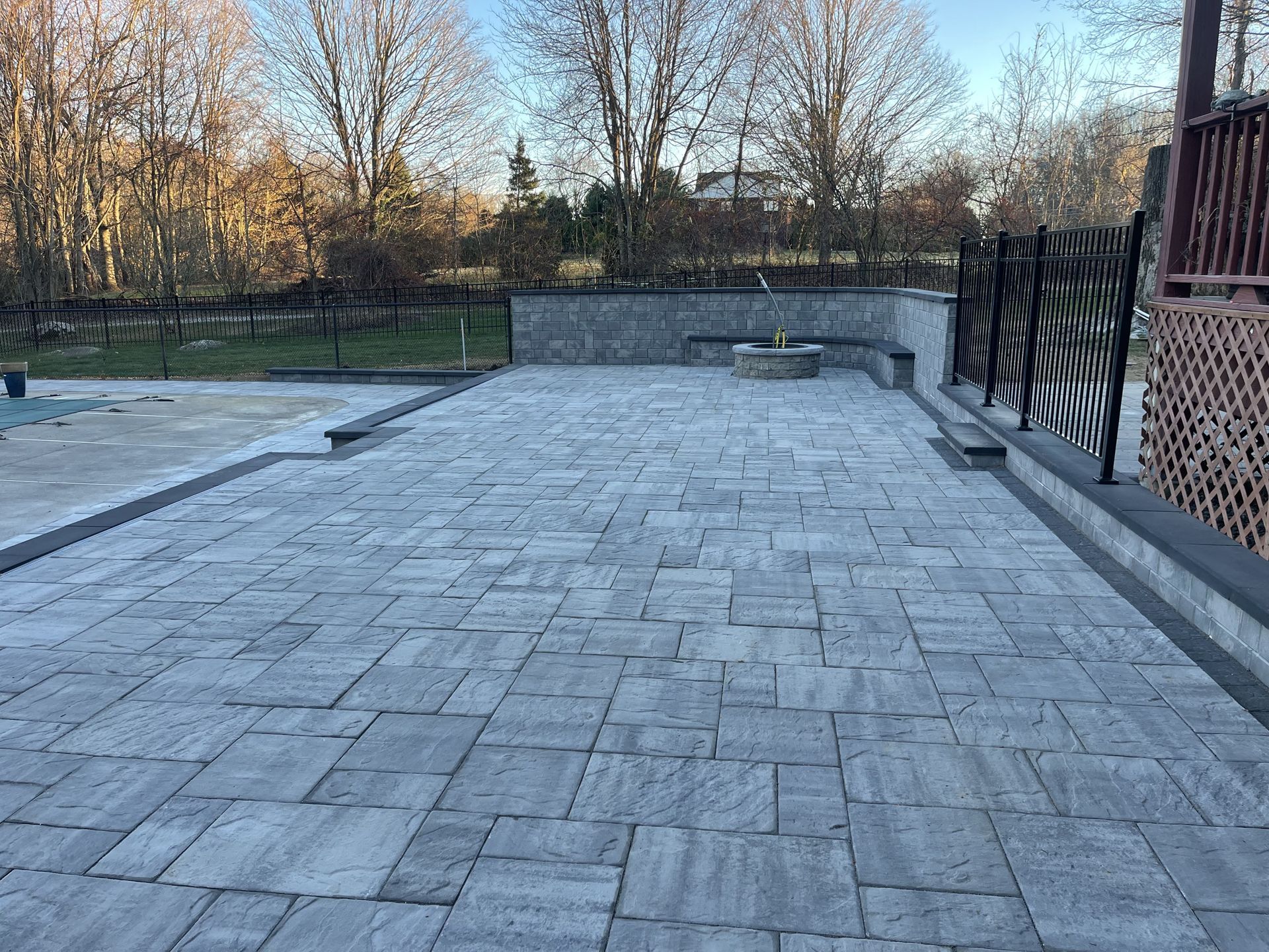 Empty stone patio with benches, black metal fence, and trees beyond a low wall