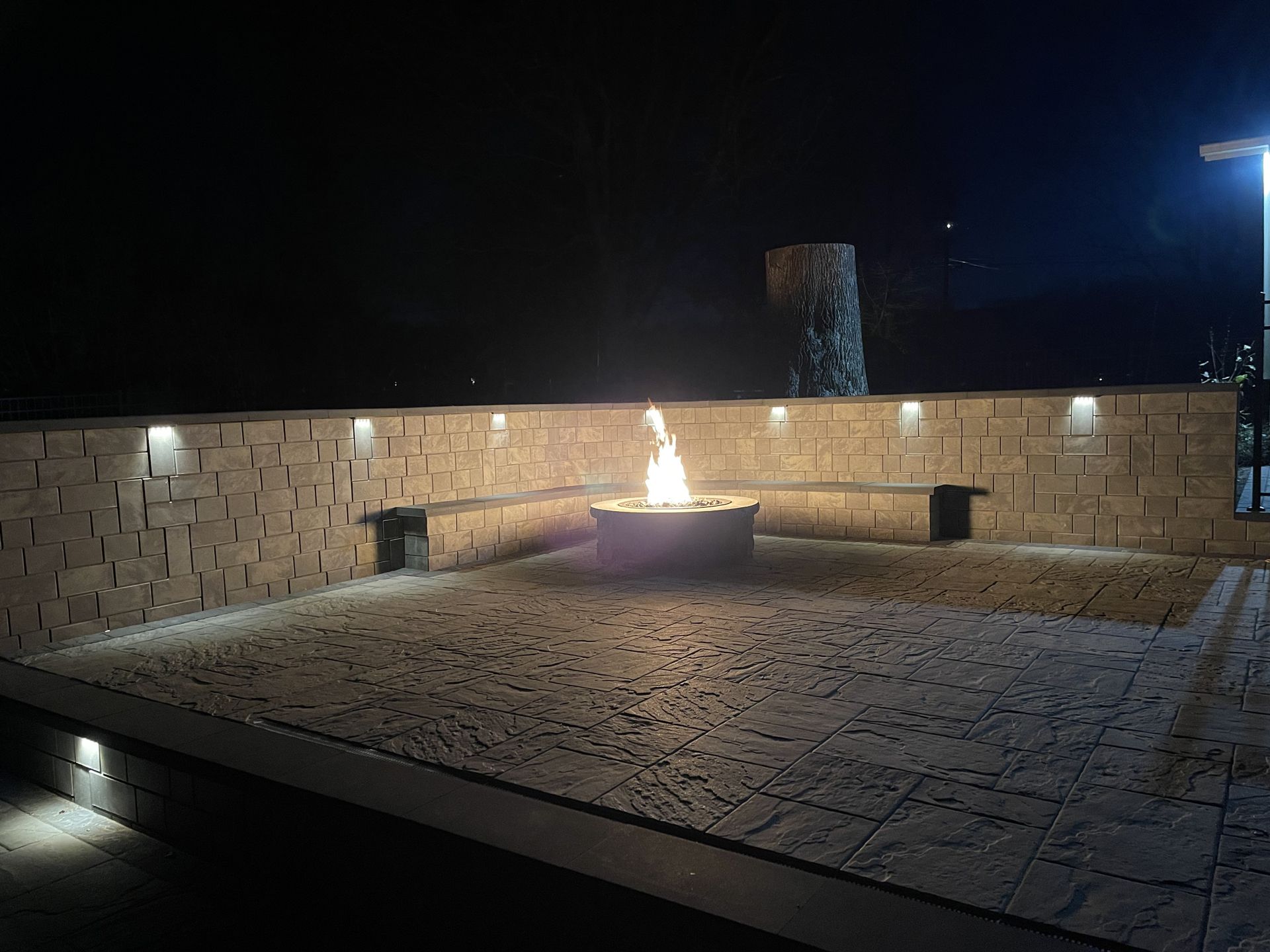 Stone courtyard at night with a small fire fountain in the center and lit wall sconces around the perimeter