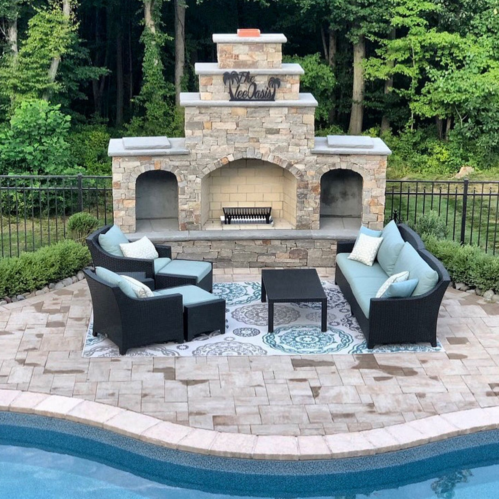 Outdoor stone fireplace with black wicker seating around a pool patio and green trees behind