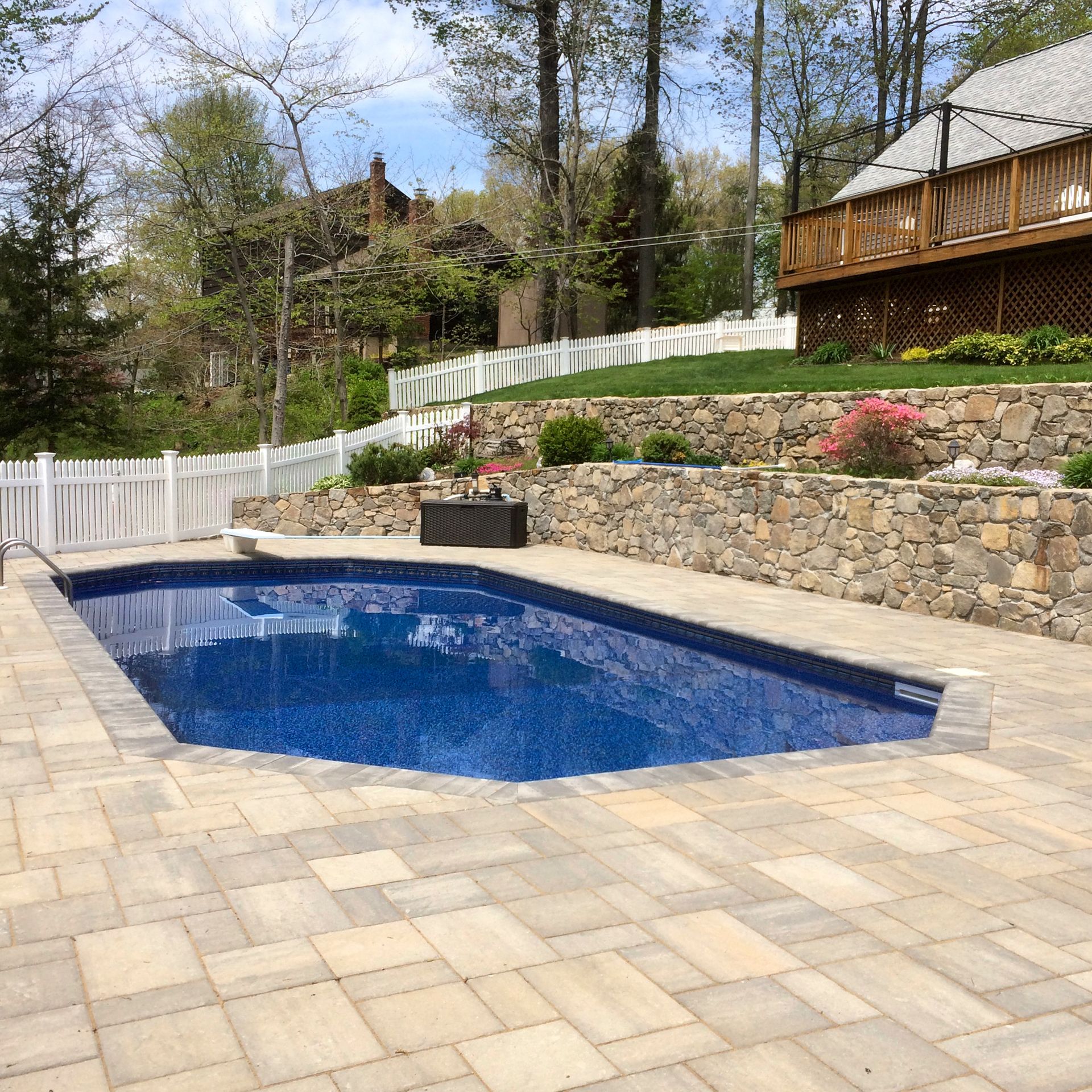 Backyard in-ground pool with blue water, stone patio, white fence, and wooded hillside