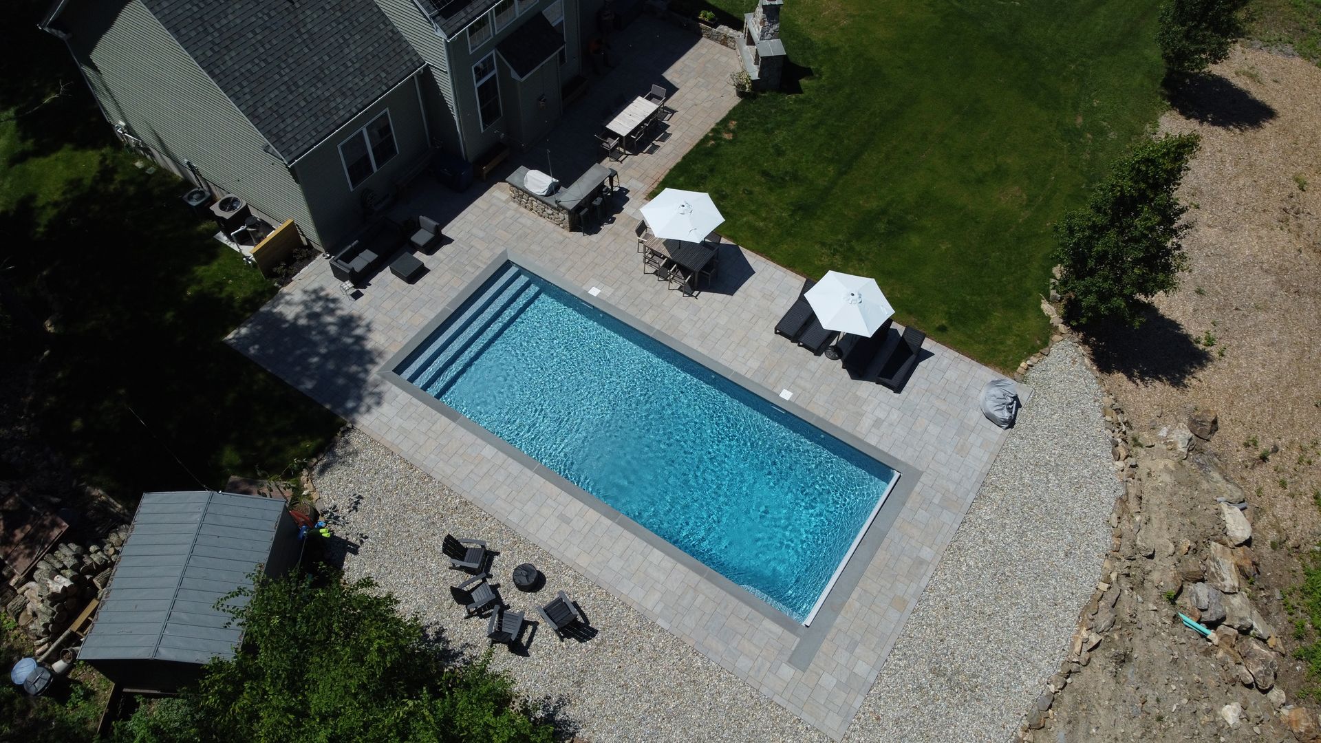 Aerial view of a backyard with a rectangular blue swimming pool, lounge chairs, and patio seating on gravel.