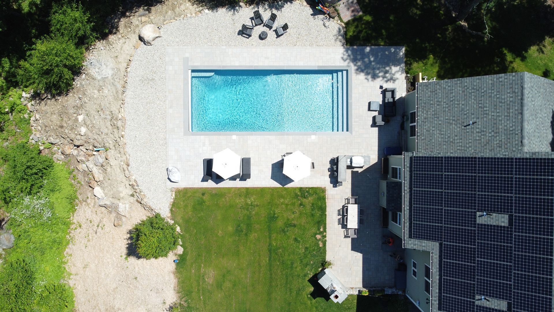 Aerial view of a backyard with a rectangular swimming pool, patio furniture, lawn, and a house roof.