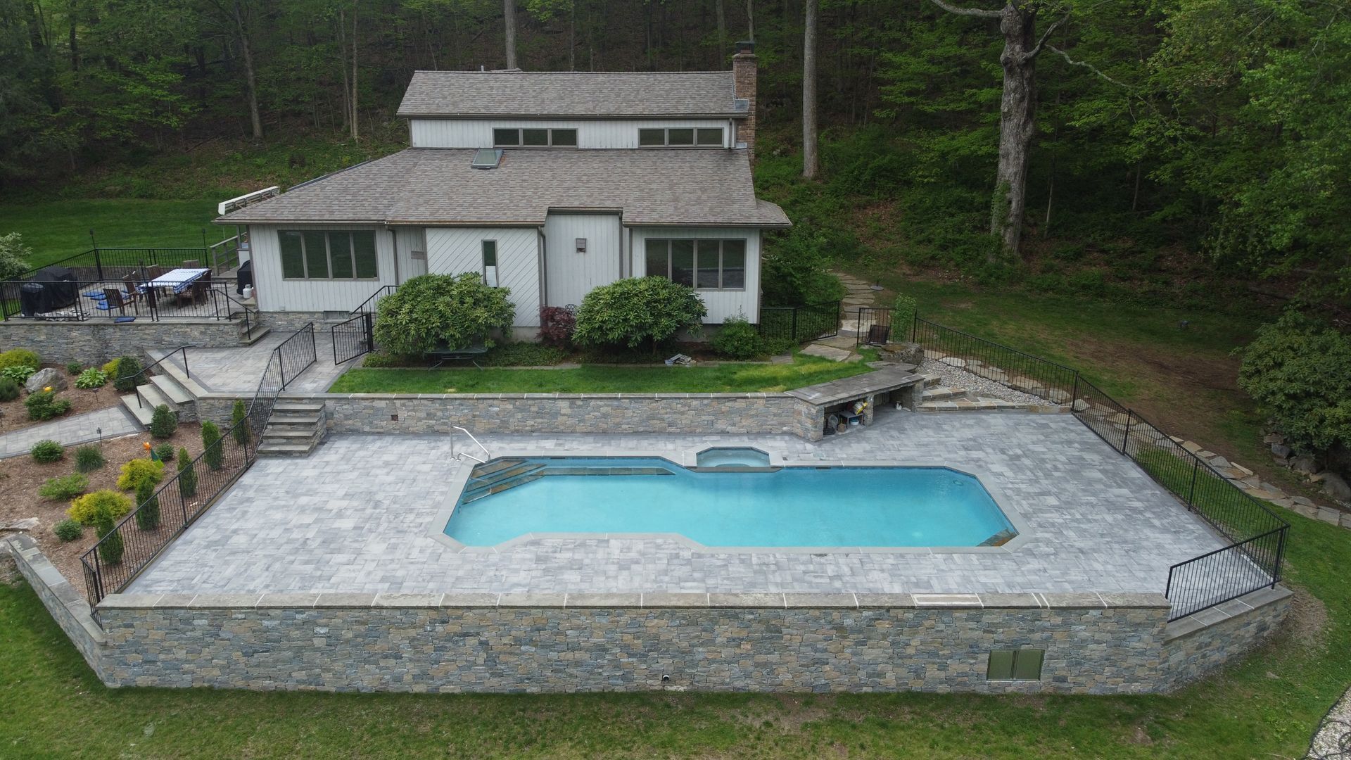 A house with a stone patio and a small turquoise swimming pool in a wooded yard.
