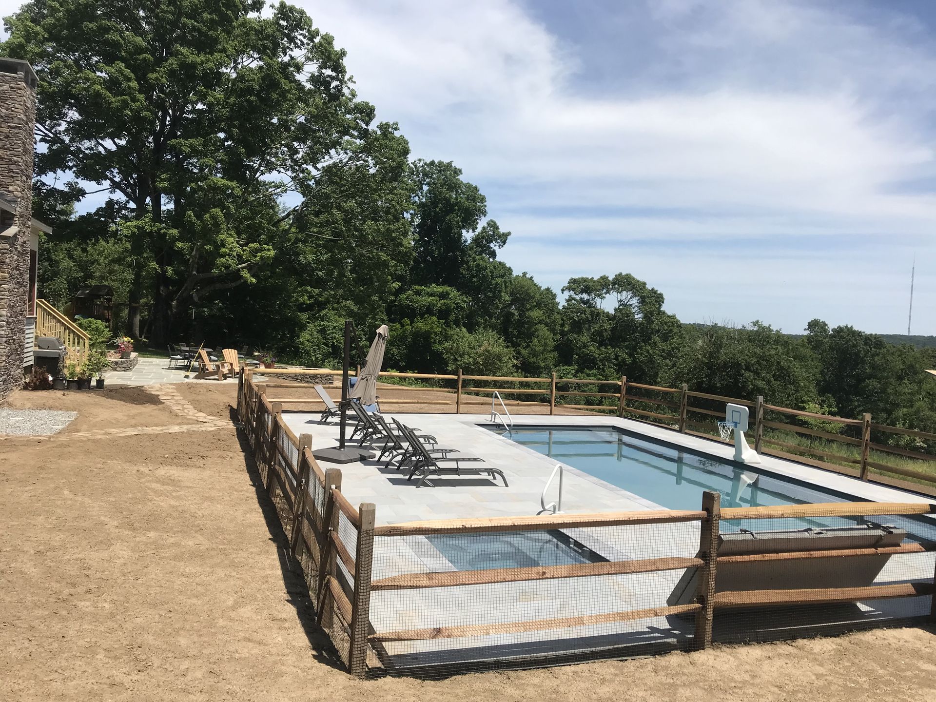 Outdoor swimming pool with lounge chairs and a fenced deck beside trees and open fields