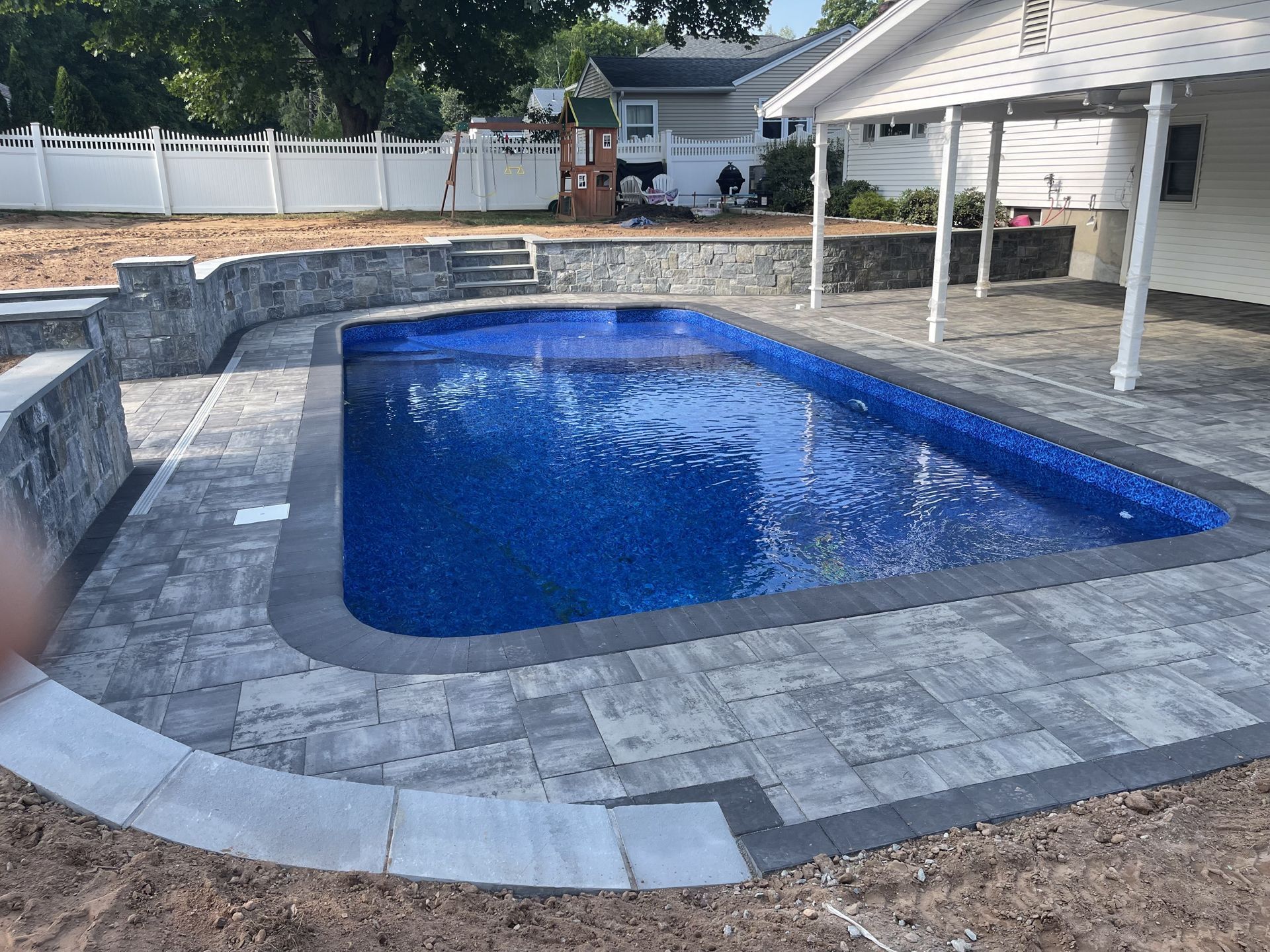 Backyard in-ground pool with blue water, gray paver deck, and white fence and patio cover nearby