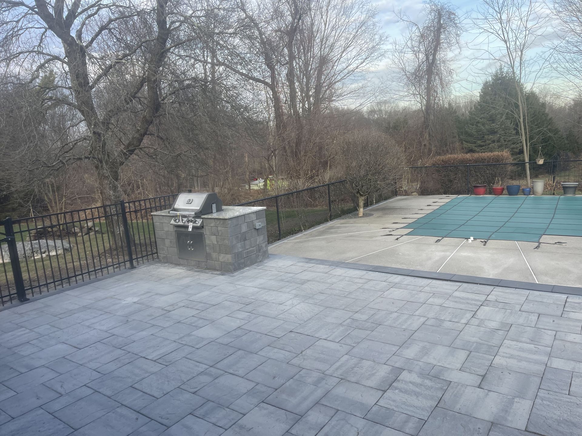 Patio with gray pavers, black fence, trees, and a covered pool area in the background
