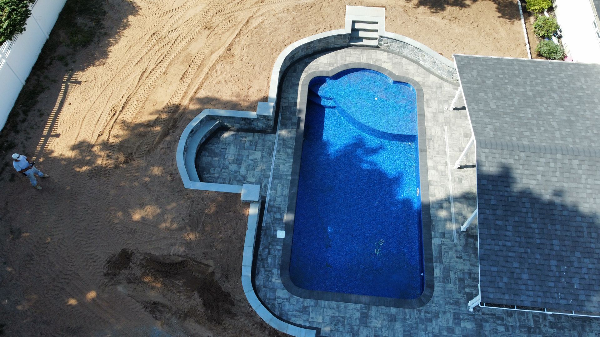 Aerial view of a backyard pool with blue water beside a patio and dirt landscaping