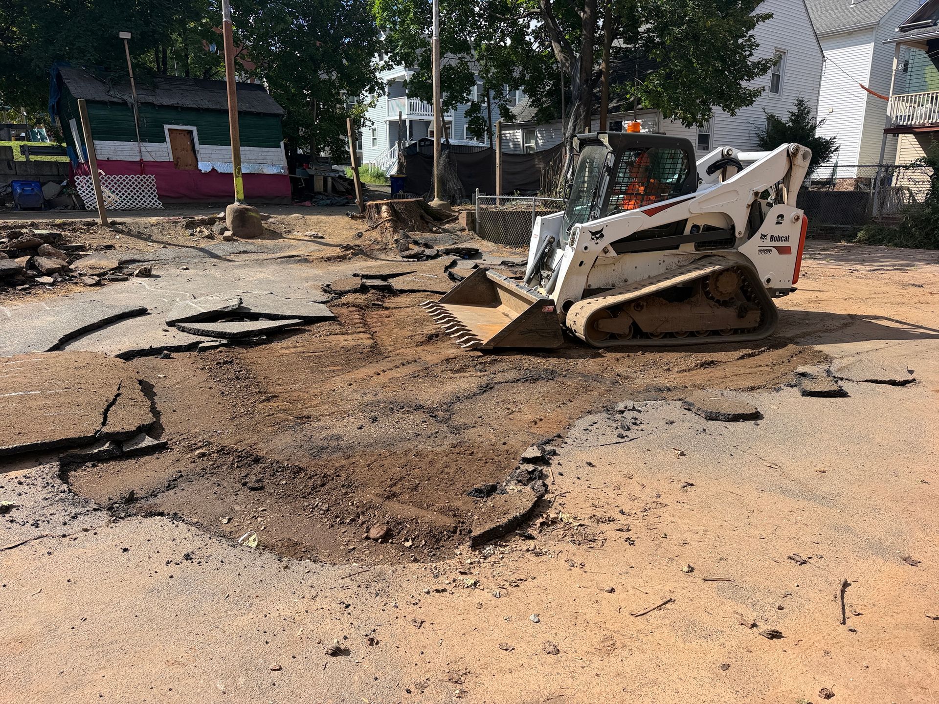 Mini excavator digging up a dirt lot with damaged asphalt near houses and parked cars