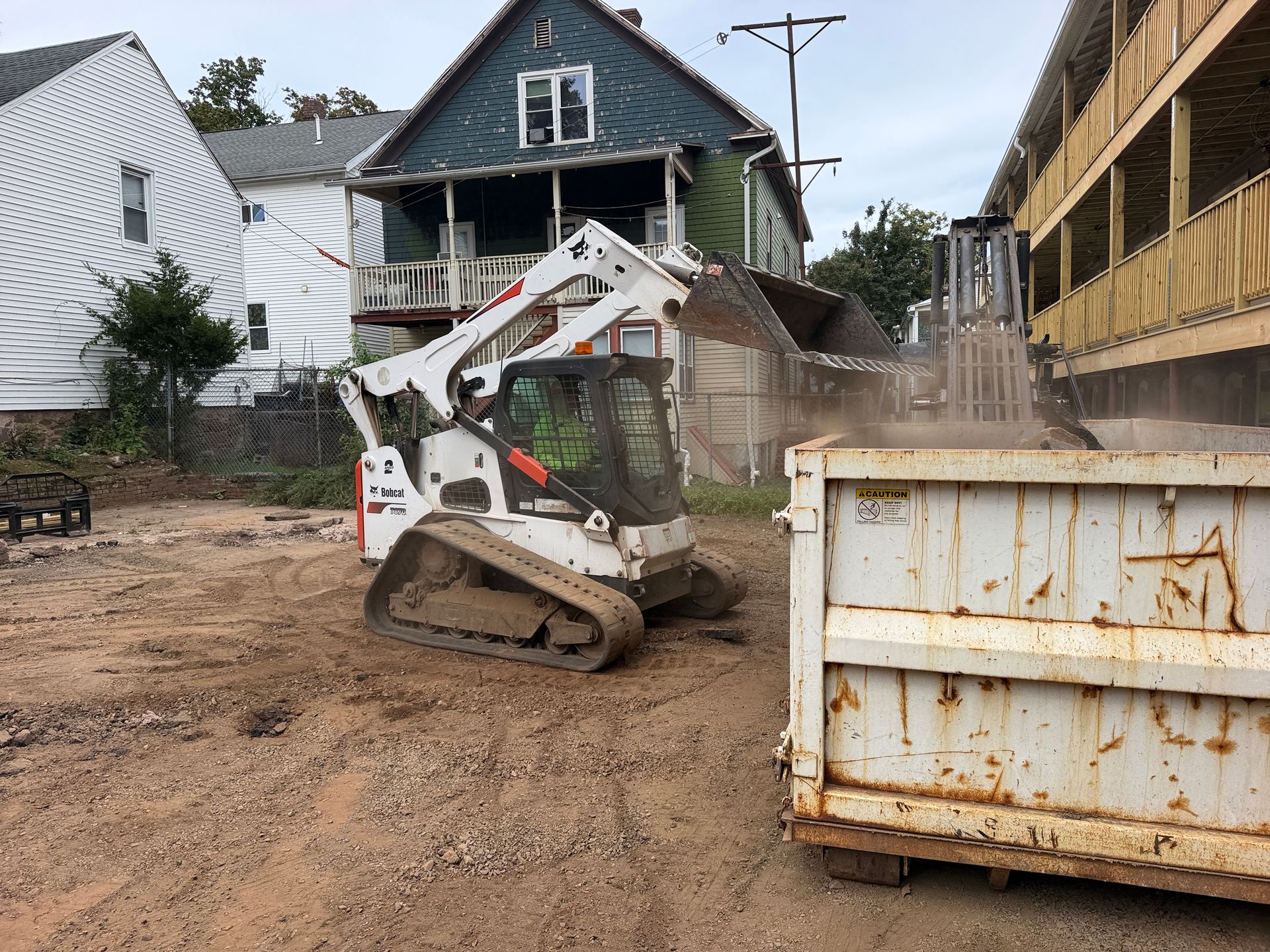 Compact loader scooping dirt at a muddy construction site beside houses and a metal dumpster