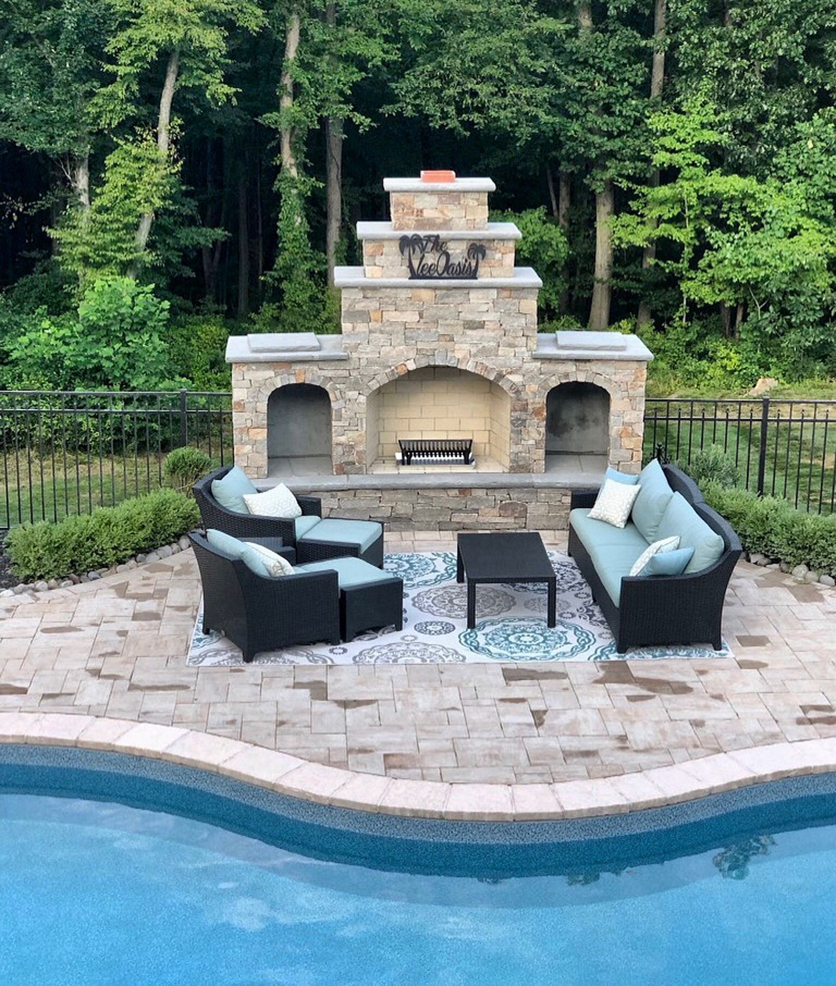 Outdoor stone fireplace with cushioned seating beside a backyard pool, framed by trees.