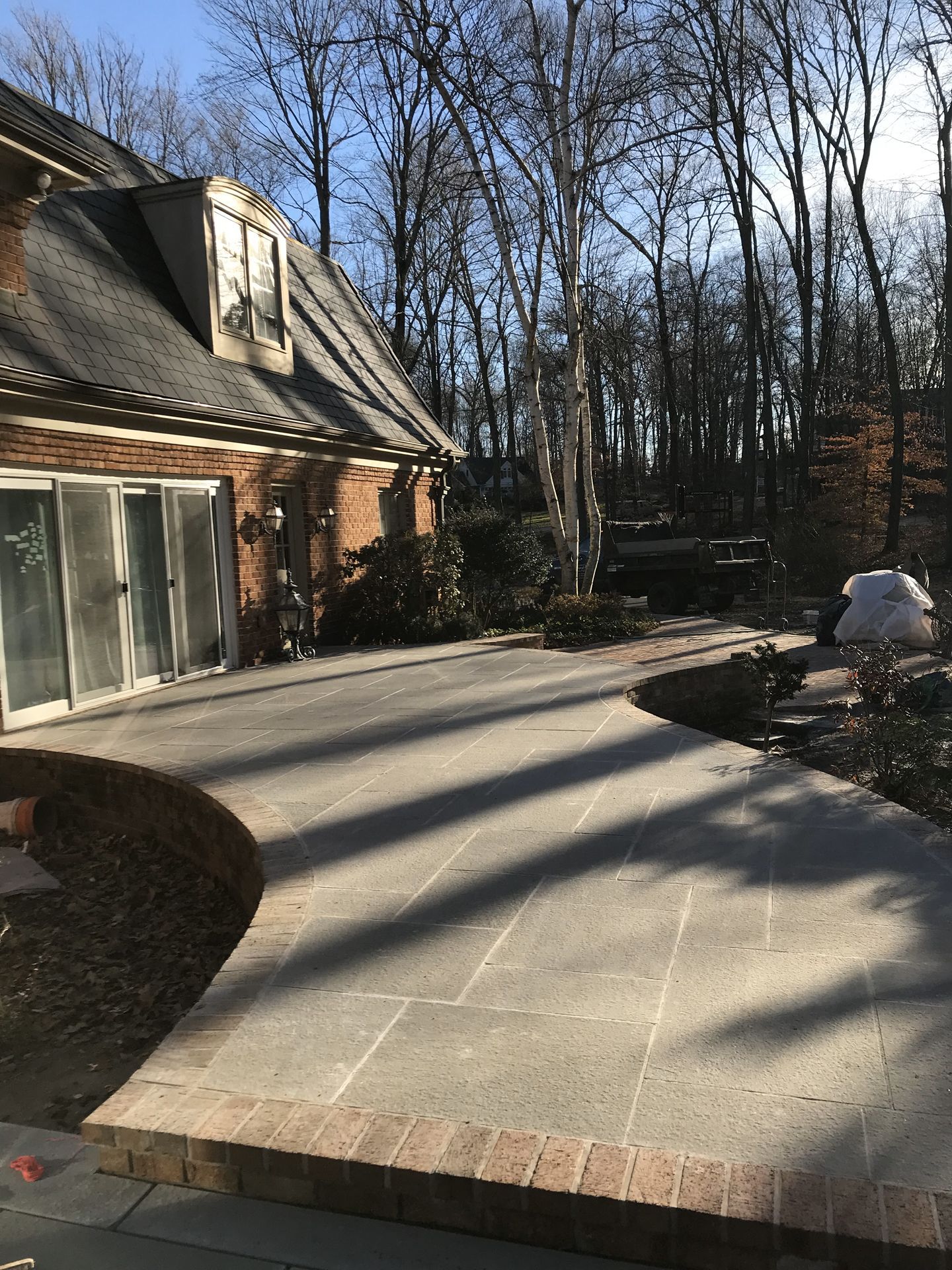 Brick patio beside a house, with curved concrete walkway and bare trees in a shaded yard