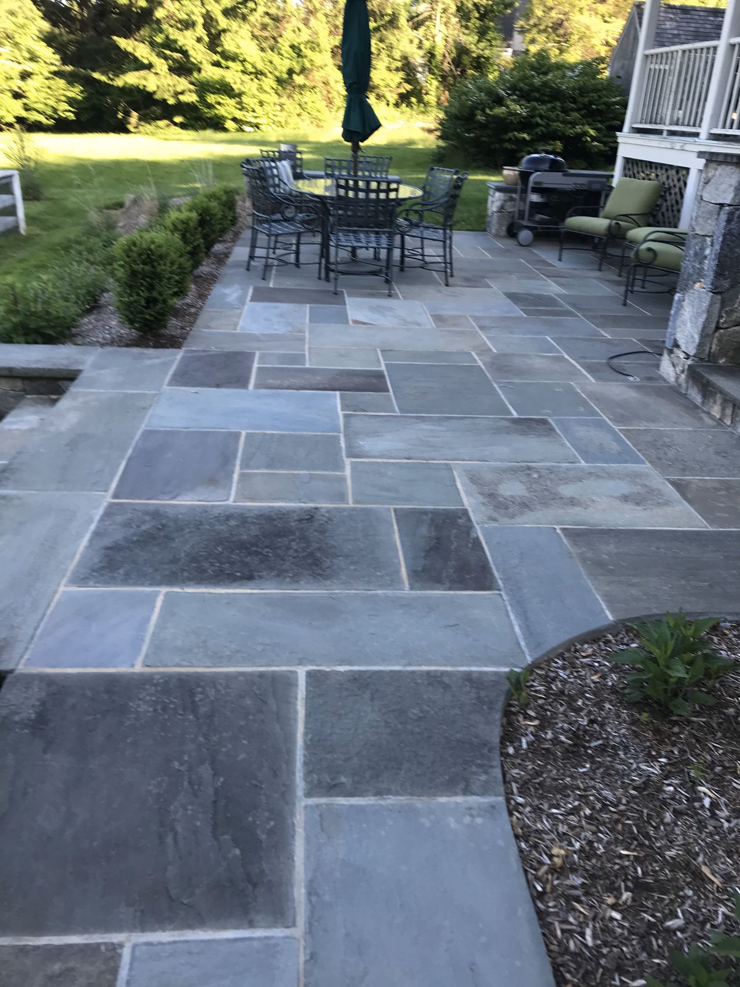 Stone patio with outdoor table, chairs, and umbrella beside a garden and house