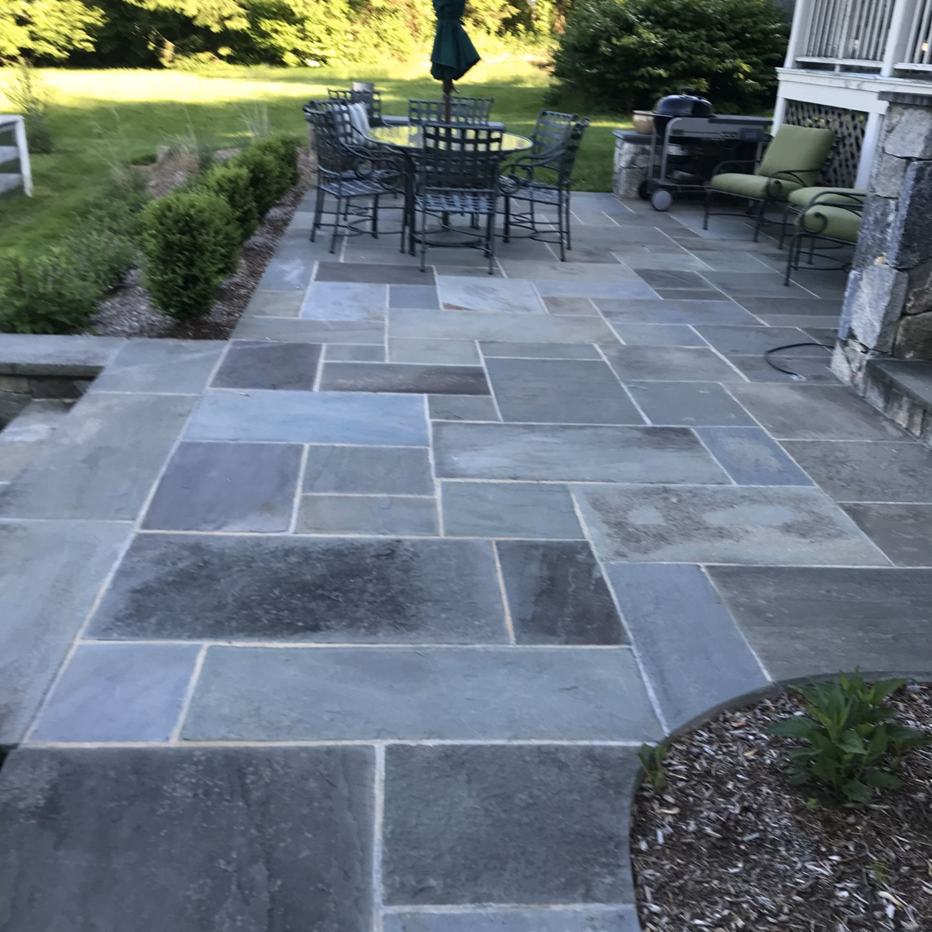 Stone patio with outdoor dining table, chairs, umbrella, and grill beside a house and garden path