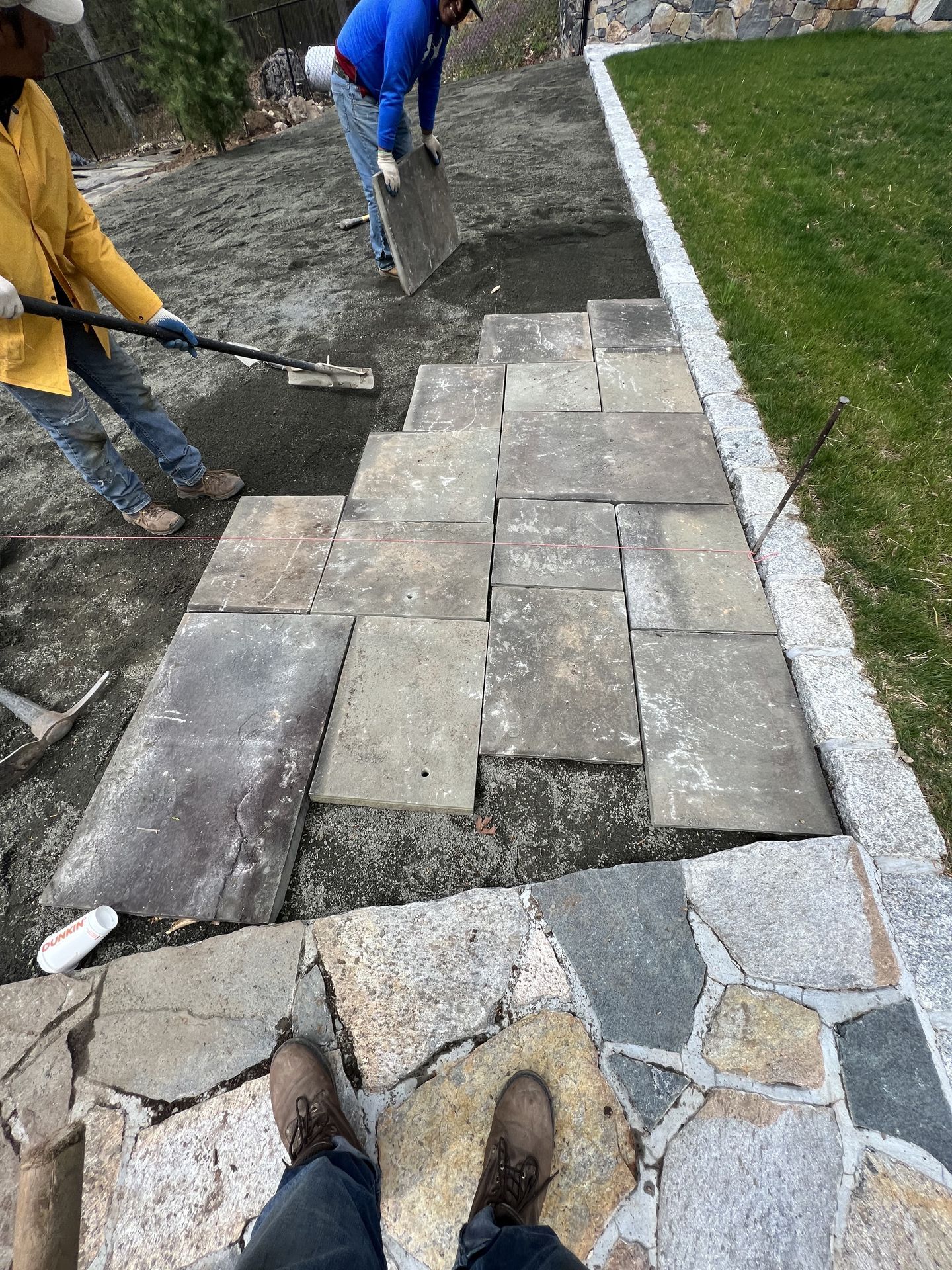 Workers laying rectangular pavers along a garden path beside a lawn and stone border