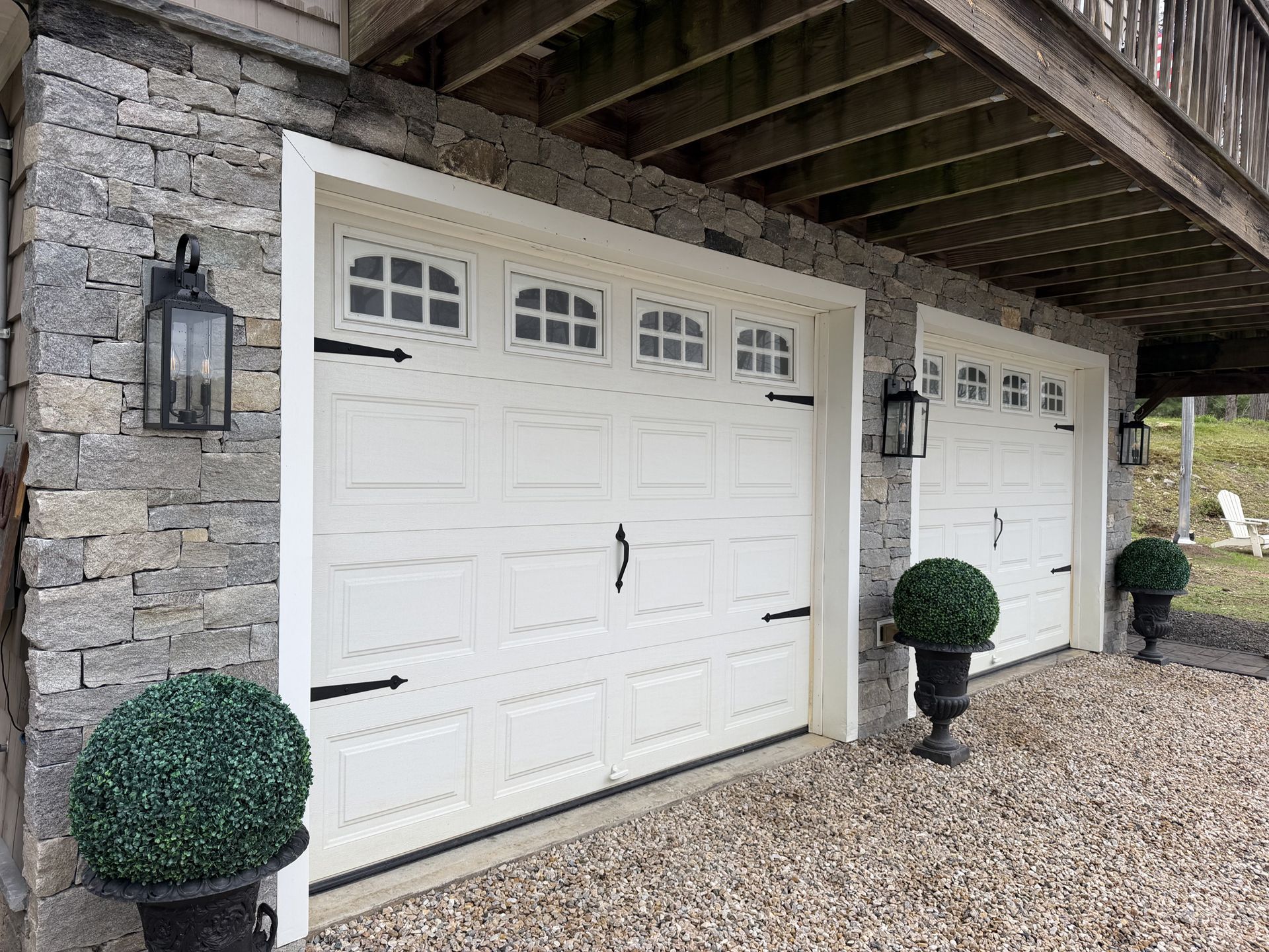 White garage doors under a stone house overhang, with trimmed topiary and gravel driveway.