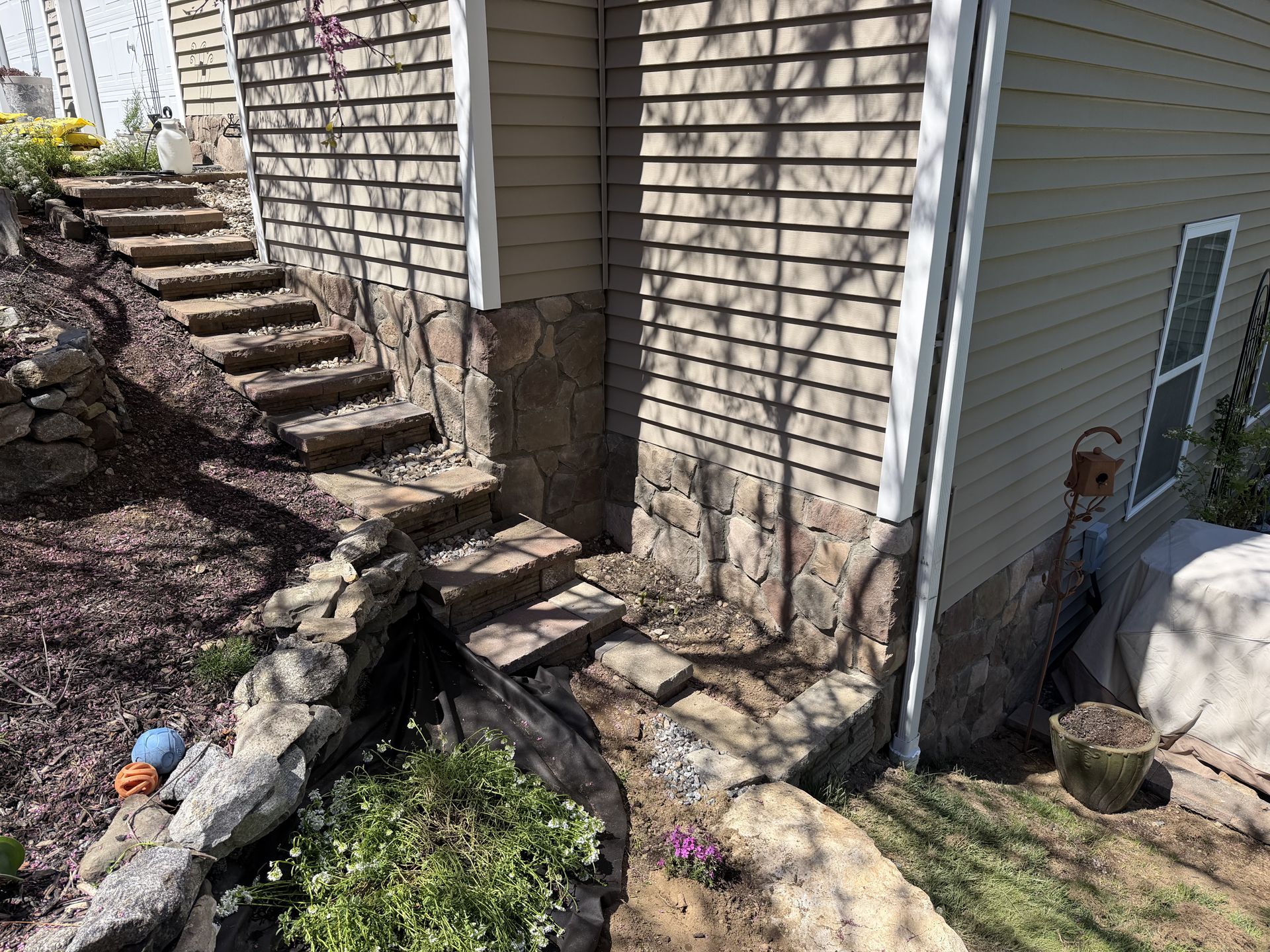 Stone steps beside a weathered house lead down to a rocky landscaped slope with sparse plants.