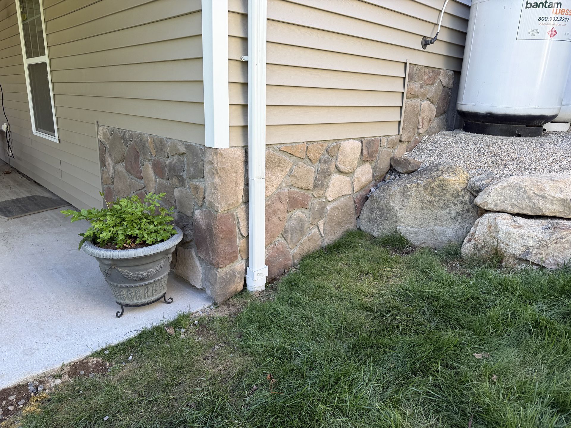 House corner with stone foundation, white downspout, potted plant on a concrete patio, and rocks by a grassy yard