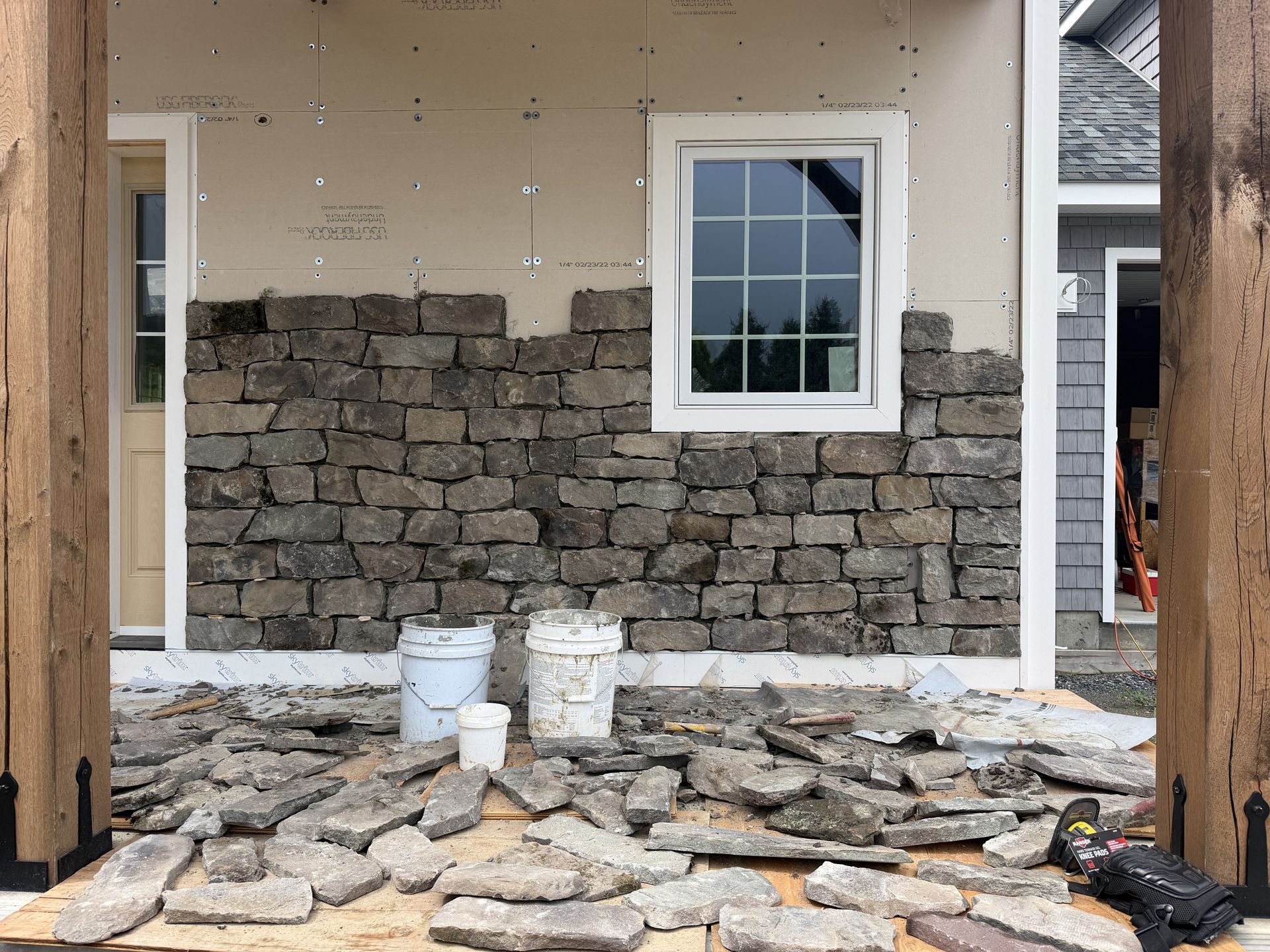 Stone veneer being installed on a house exterior, with tools and materials on the ground.