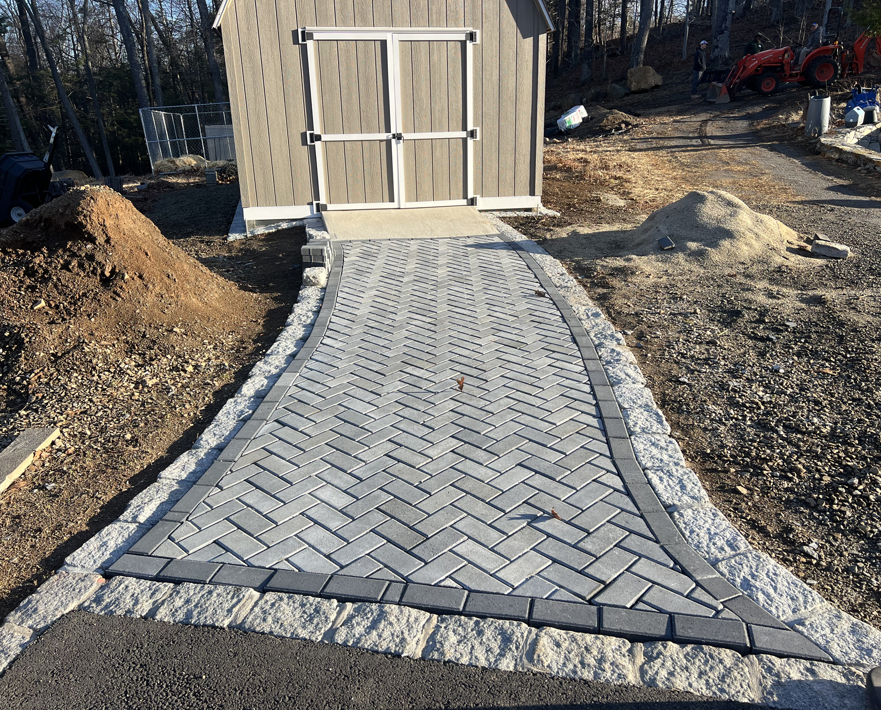 Gray herringbone paver walkway leading to a wooden double gate, bordered by mulch and gravel
