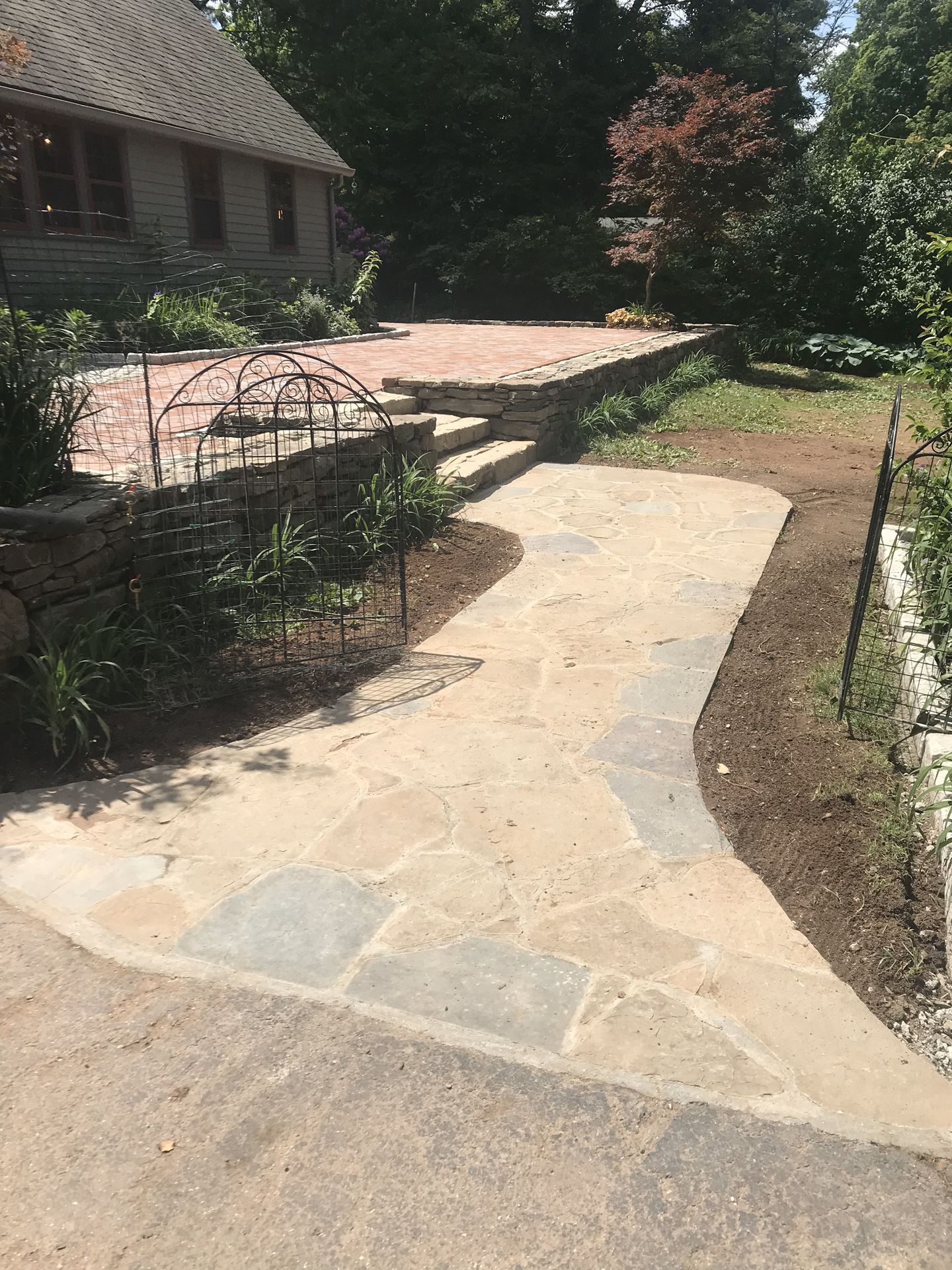 Stone walkway curving through a garden beside a house, with brick patio and greenery.