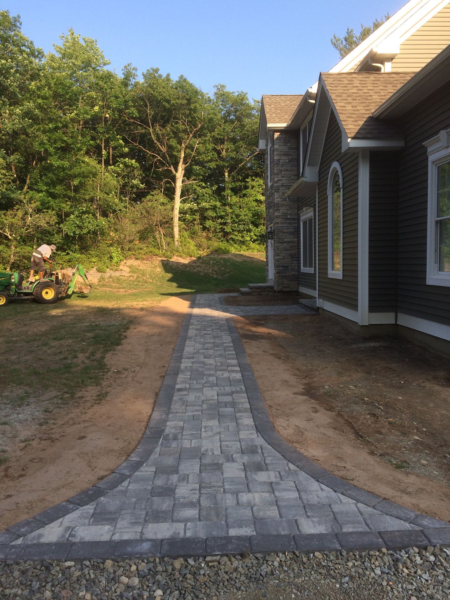 Curved gray paver walkway leading to a house, with dirt sides and green trees in the background