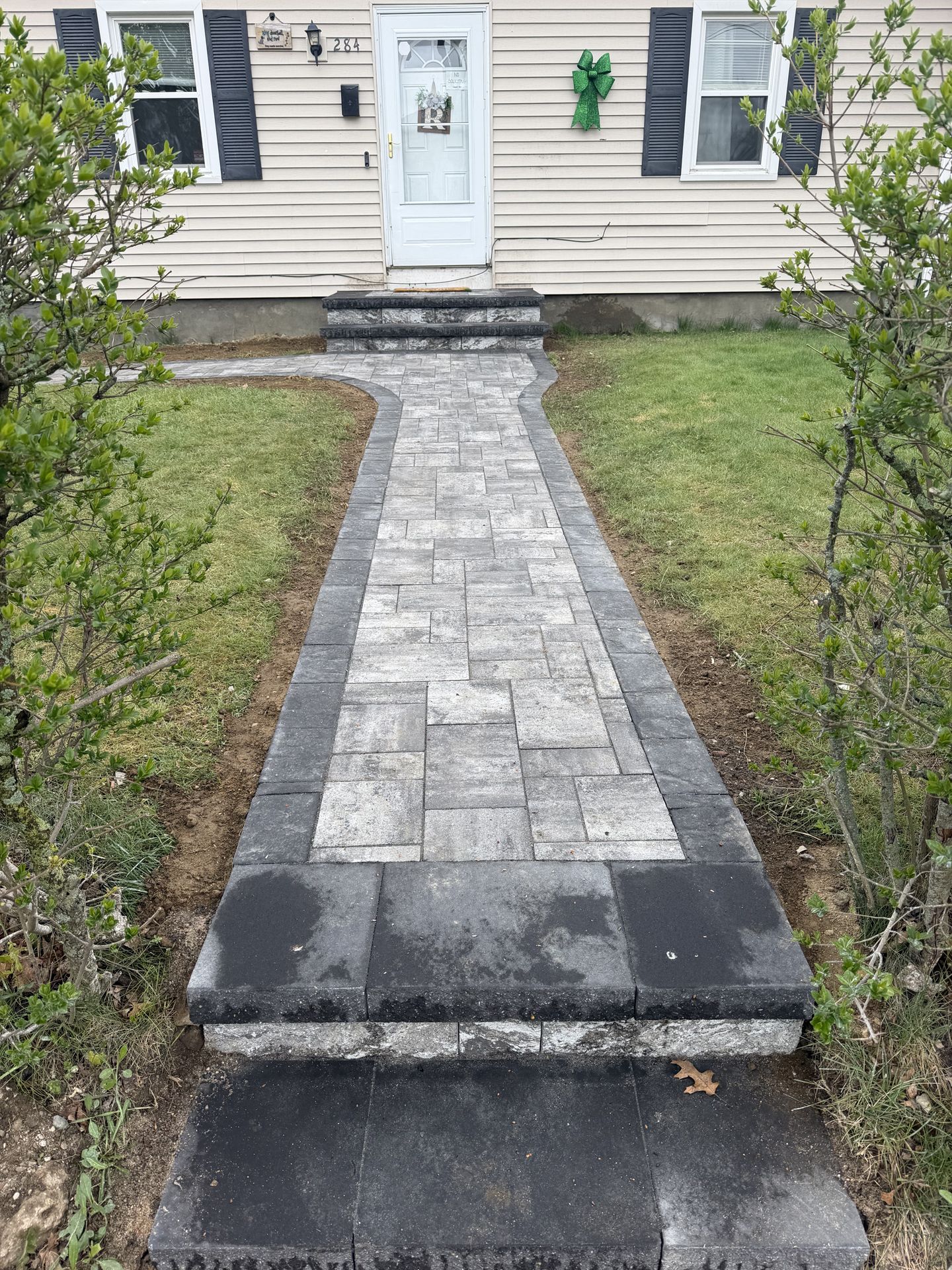 Front walkway of gray stone pavers leading to a white house door, bordered by grass and shrubs.