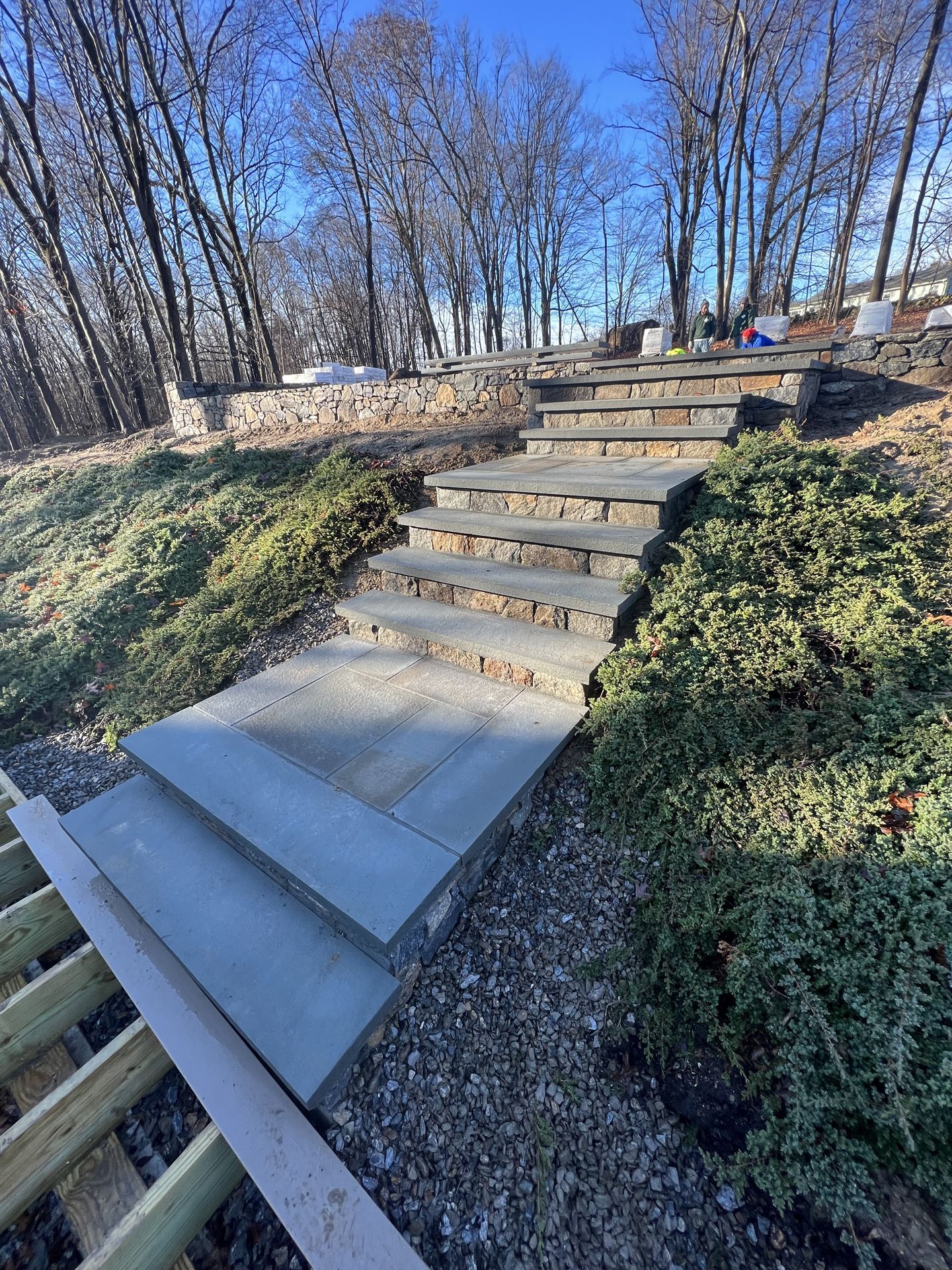 Steep outdoor wooden stairs on a wooded hillside with gravel and low shrubs under a clear sky