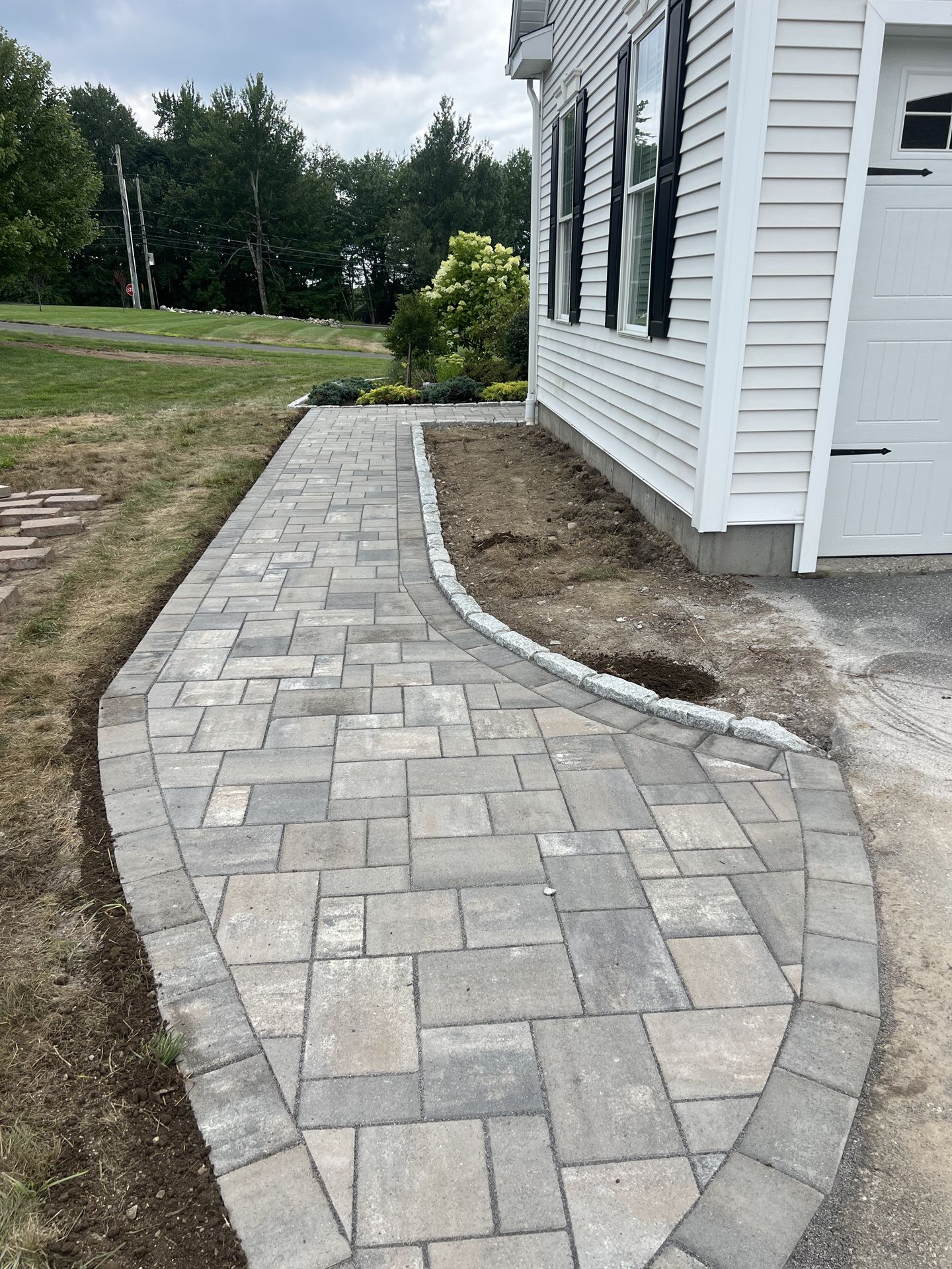 Curved paver walkway beside a white house, bordered by gravel and grass.