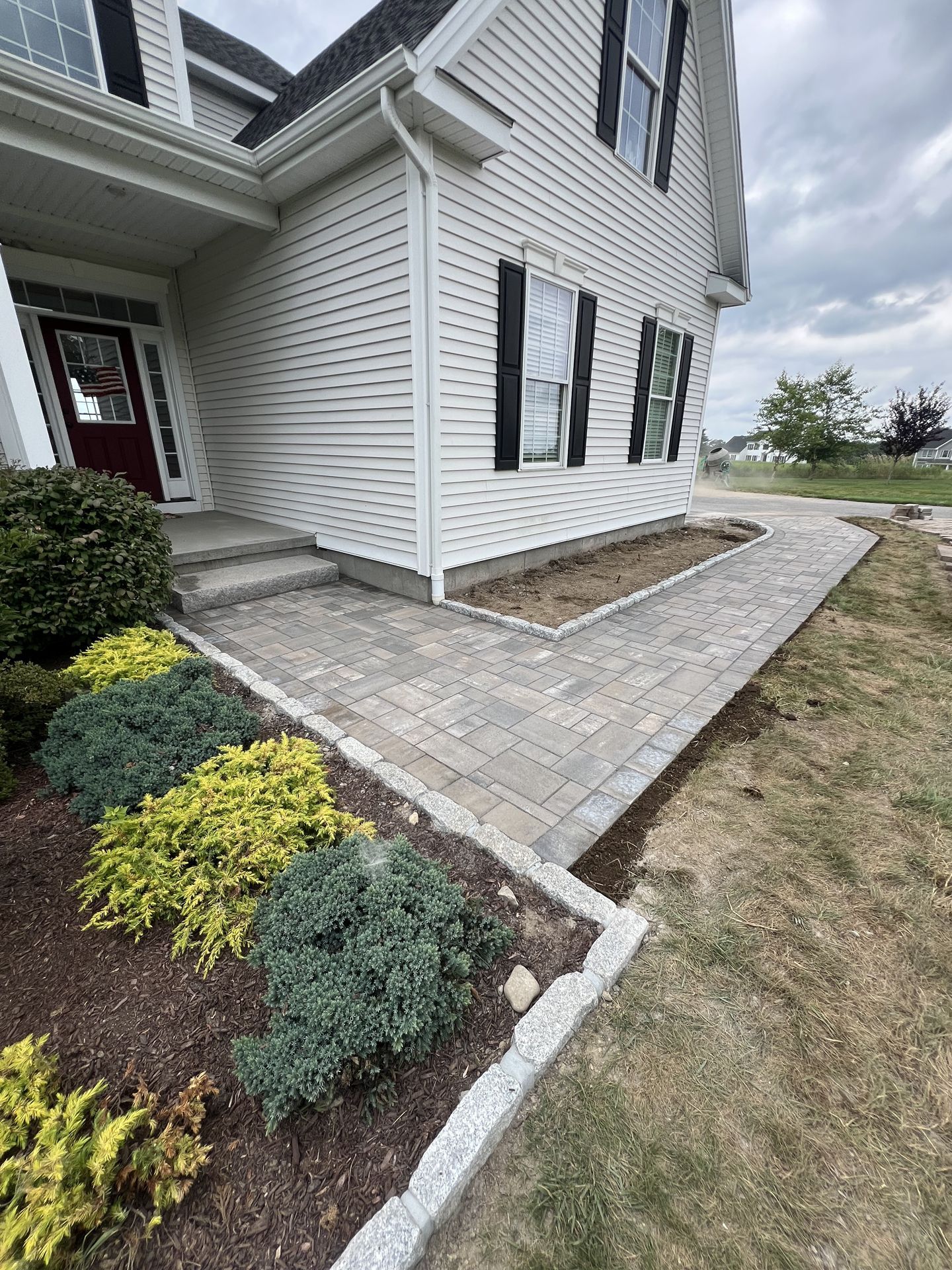 House entrance with paver walkway, white siding, black shutters, and landscaped shrubs beside a lawn