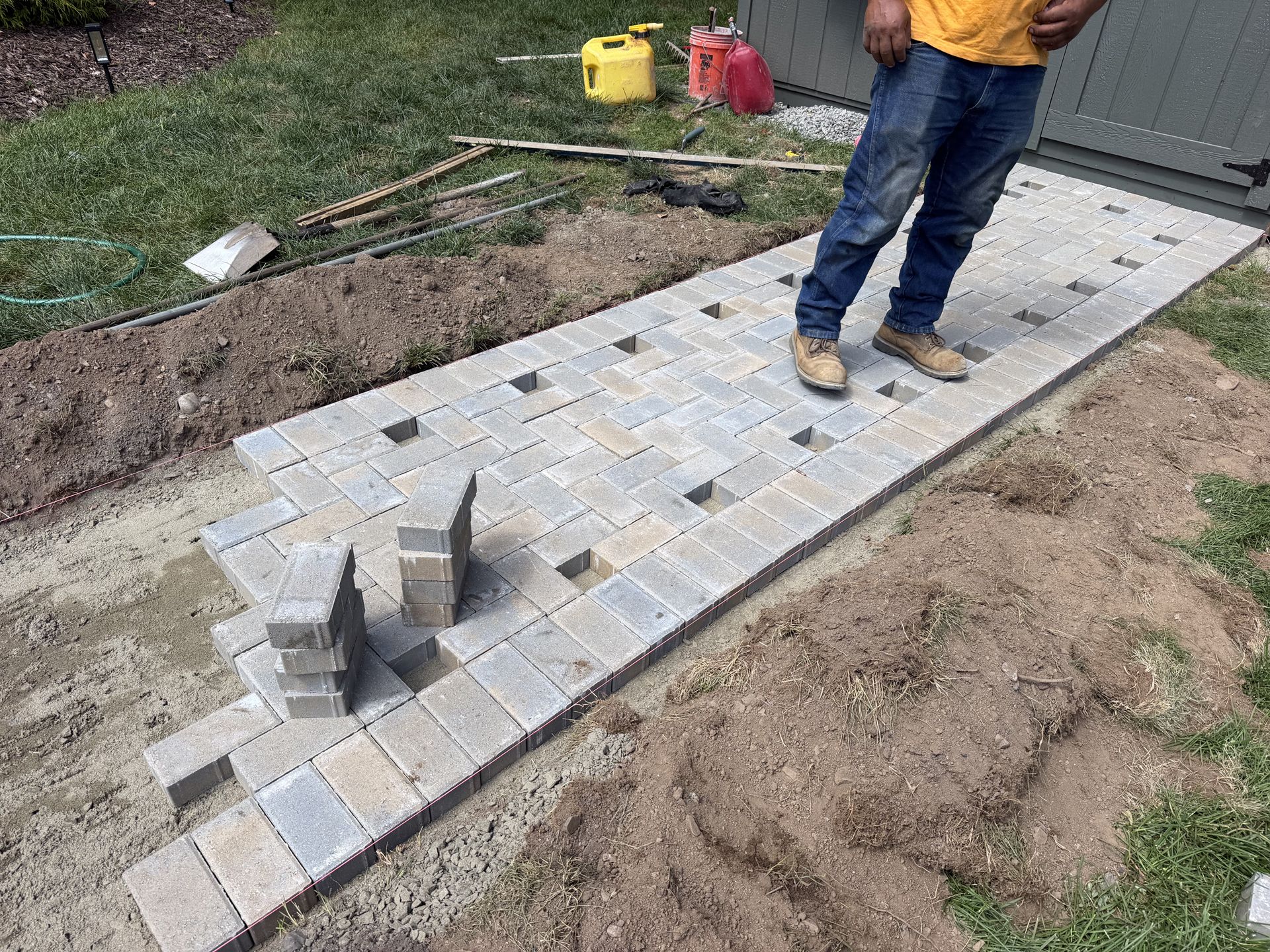 Partially paved walkway of gray concrete pavers with a worker standing beside it in a yard
