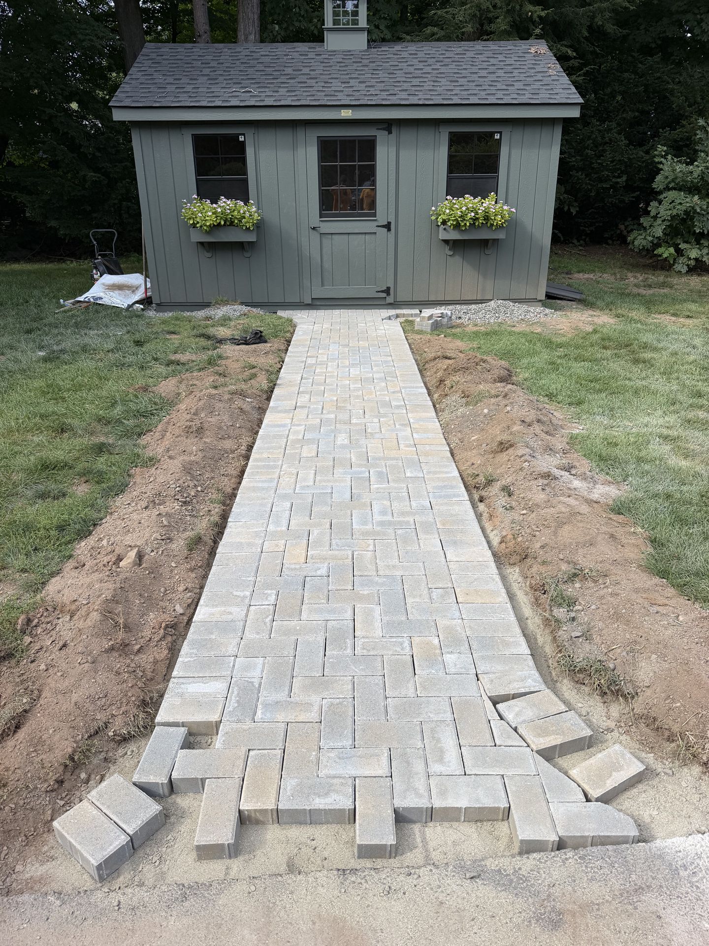 Stone walkway under construction leading to a small gray shed with flower boxes