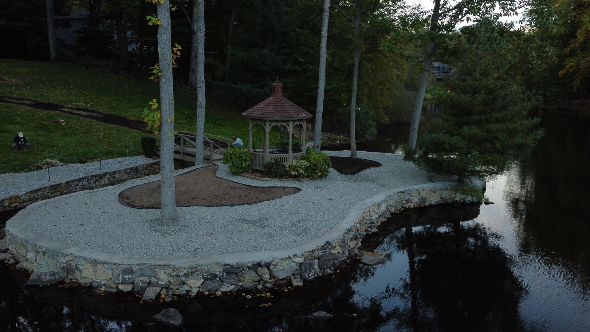 Small stone gazebo by a dark pond, surrounded by trees and a curved gravel path at dusk
