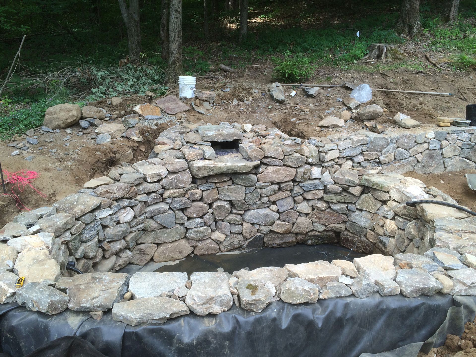 Stone-lined pond under construction in a wooded area, with black liner and stacked rocks.