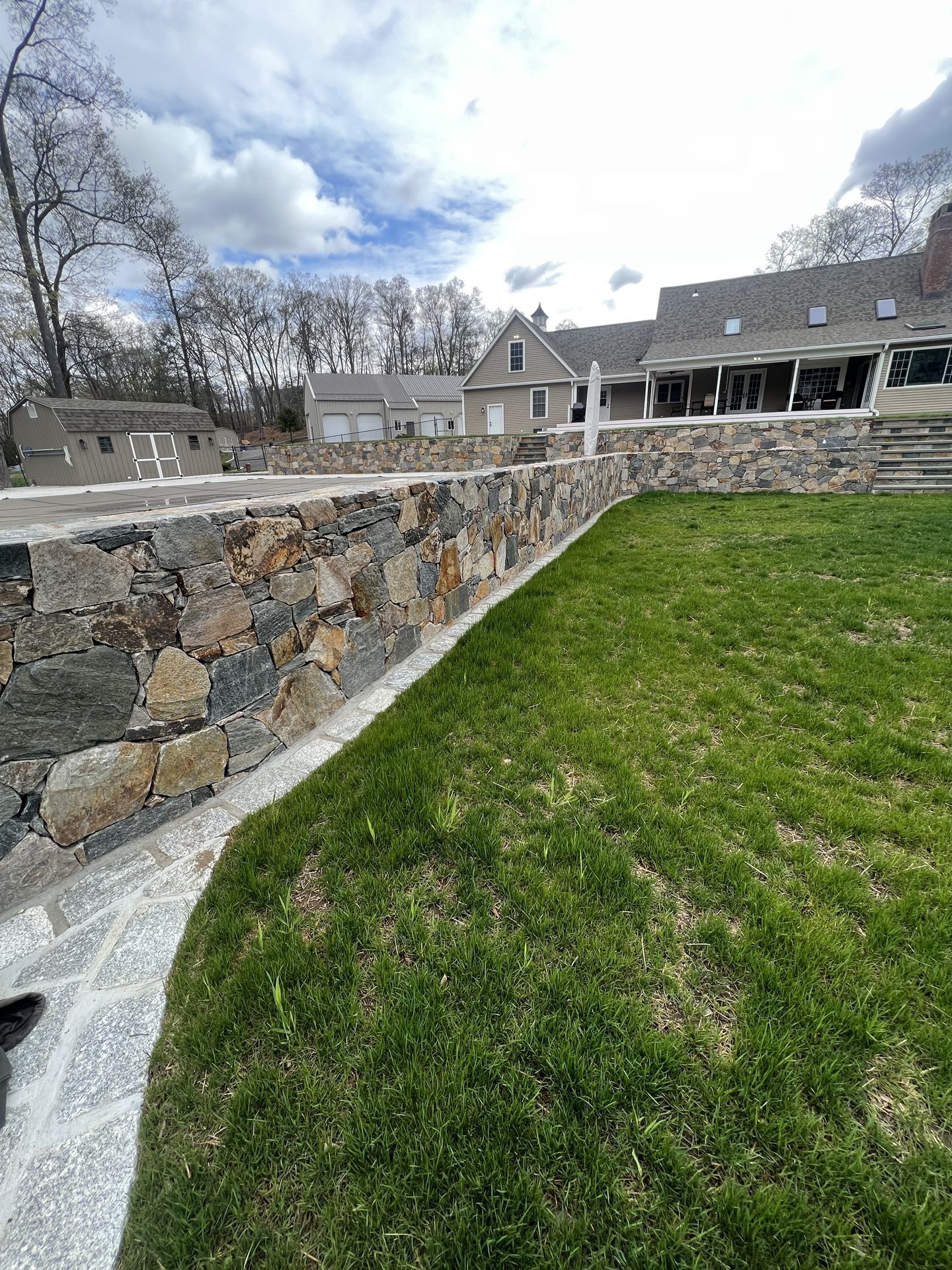 Stone retaining wall beside a grassy yard in front of a stone house under a cloudy sky.