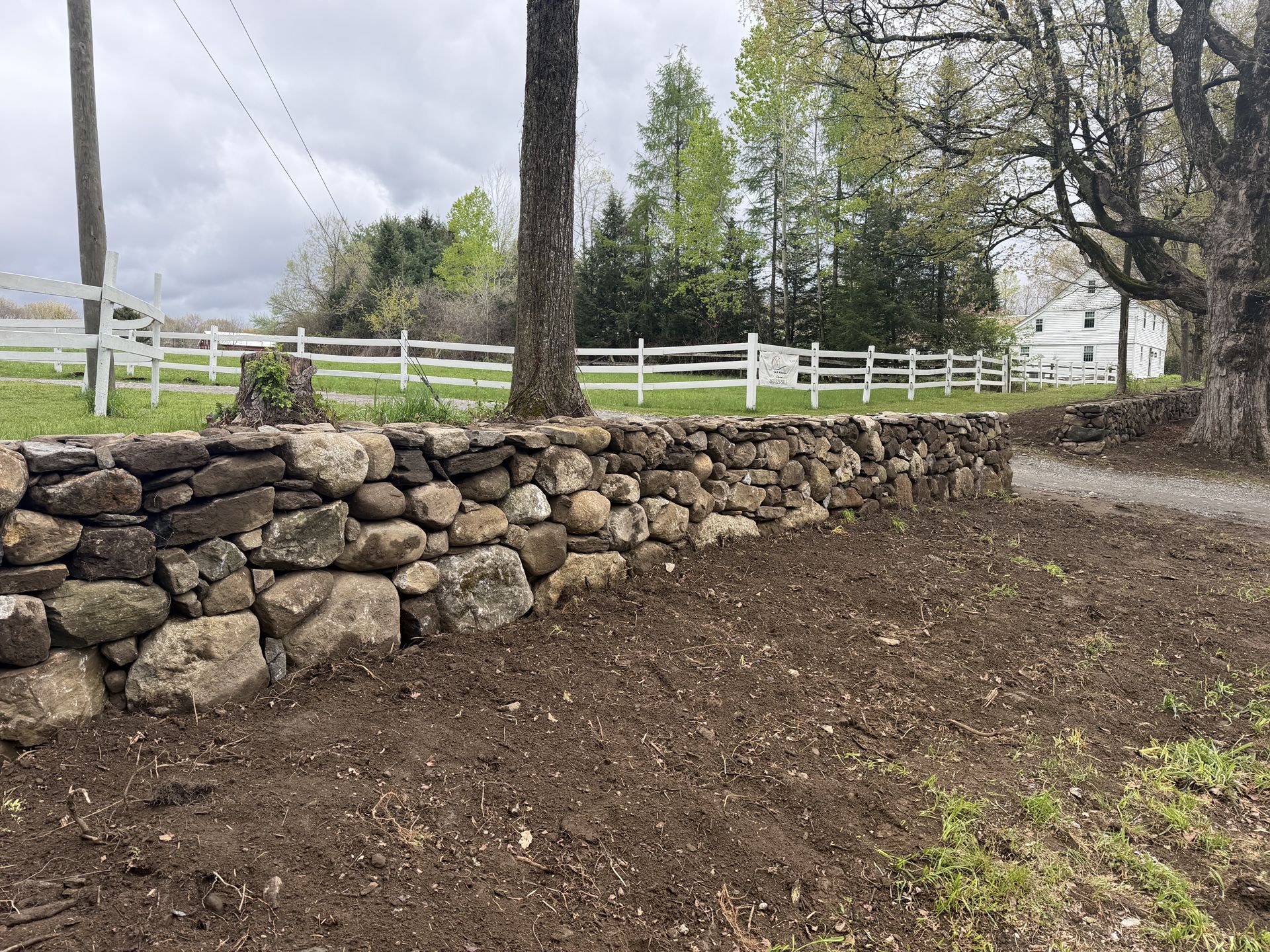 Stone retaining wall beside a dirt path, with white fence and trees in a grassy yard