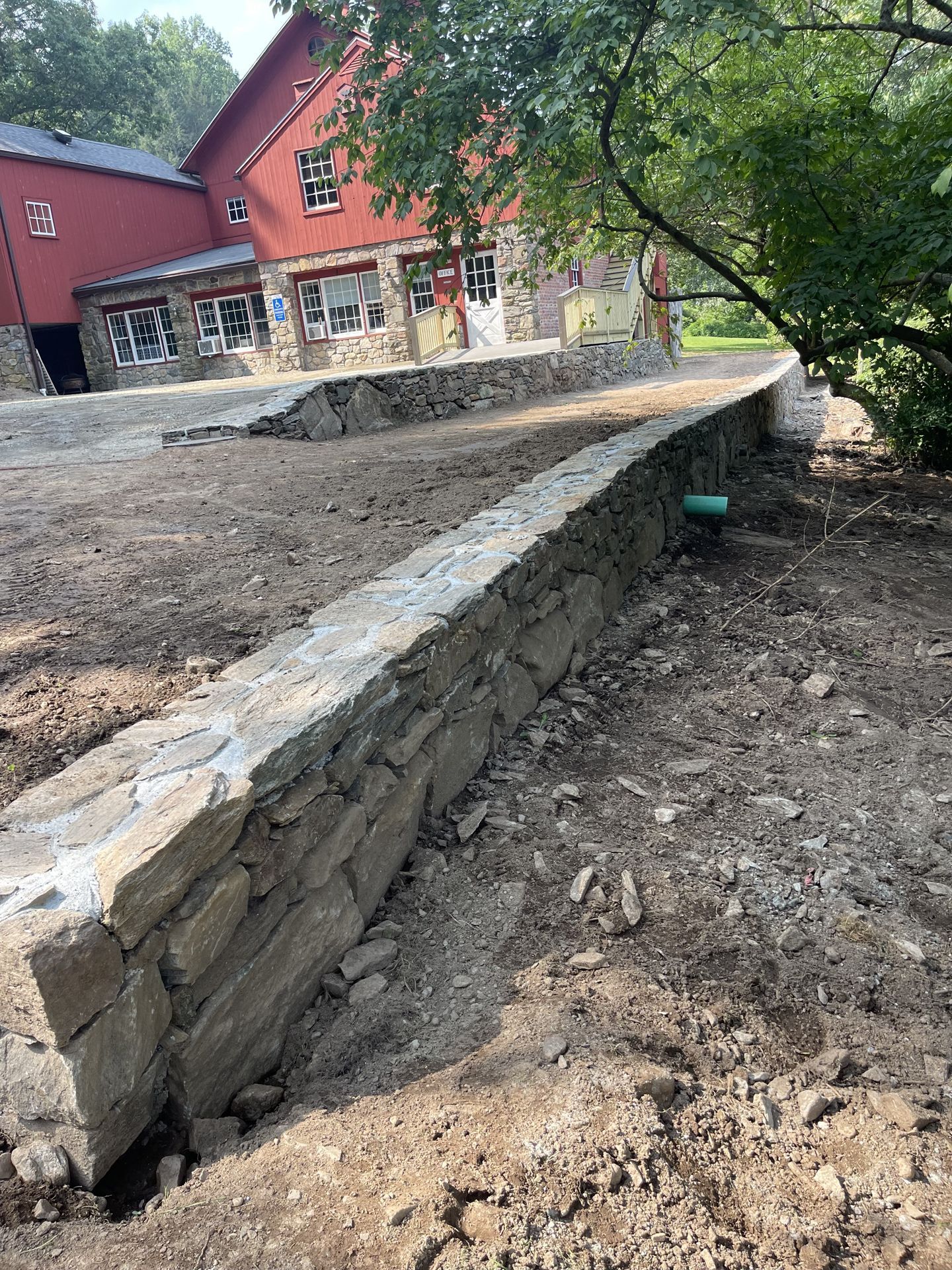 Stone retaining wall beside a dirt path, with a red building and trees in the background