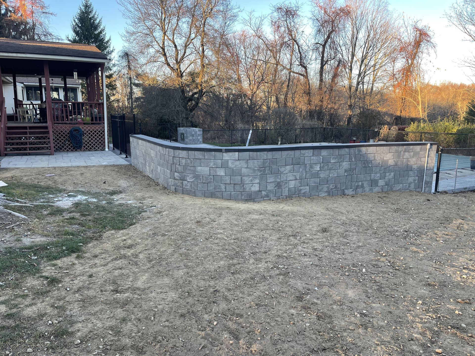 Backyard with a stone retaining wall, dry grass, and a porch beside leafless trees at sunset