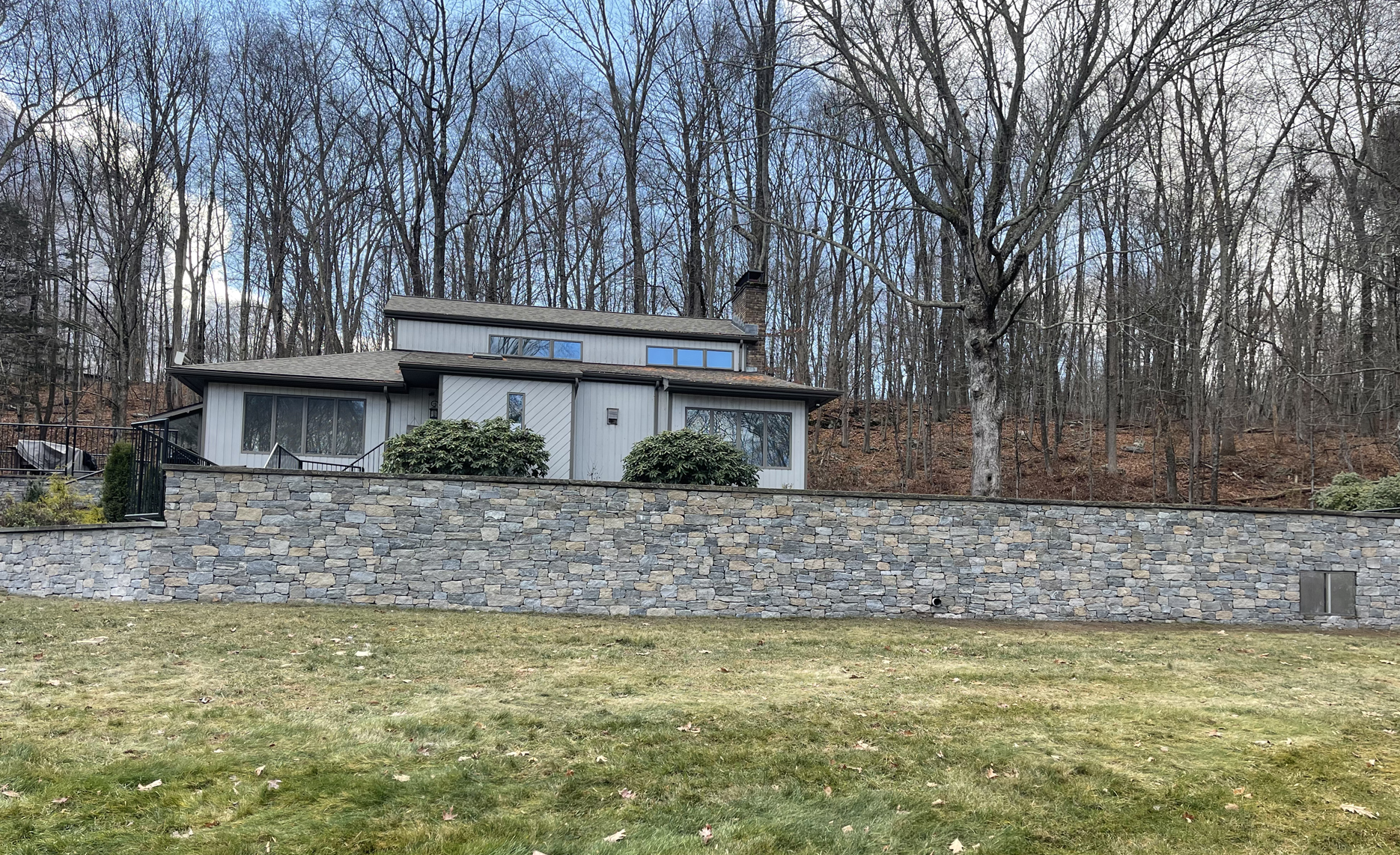 Small white building behind a stone retaining wall in a wooded area under overcast sky