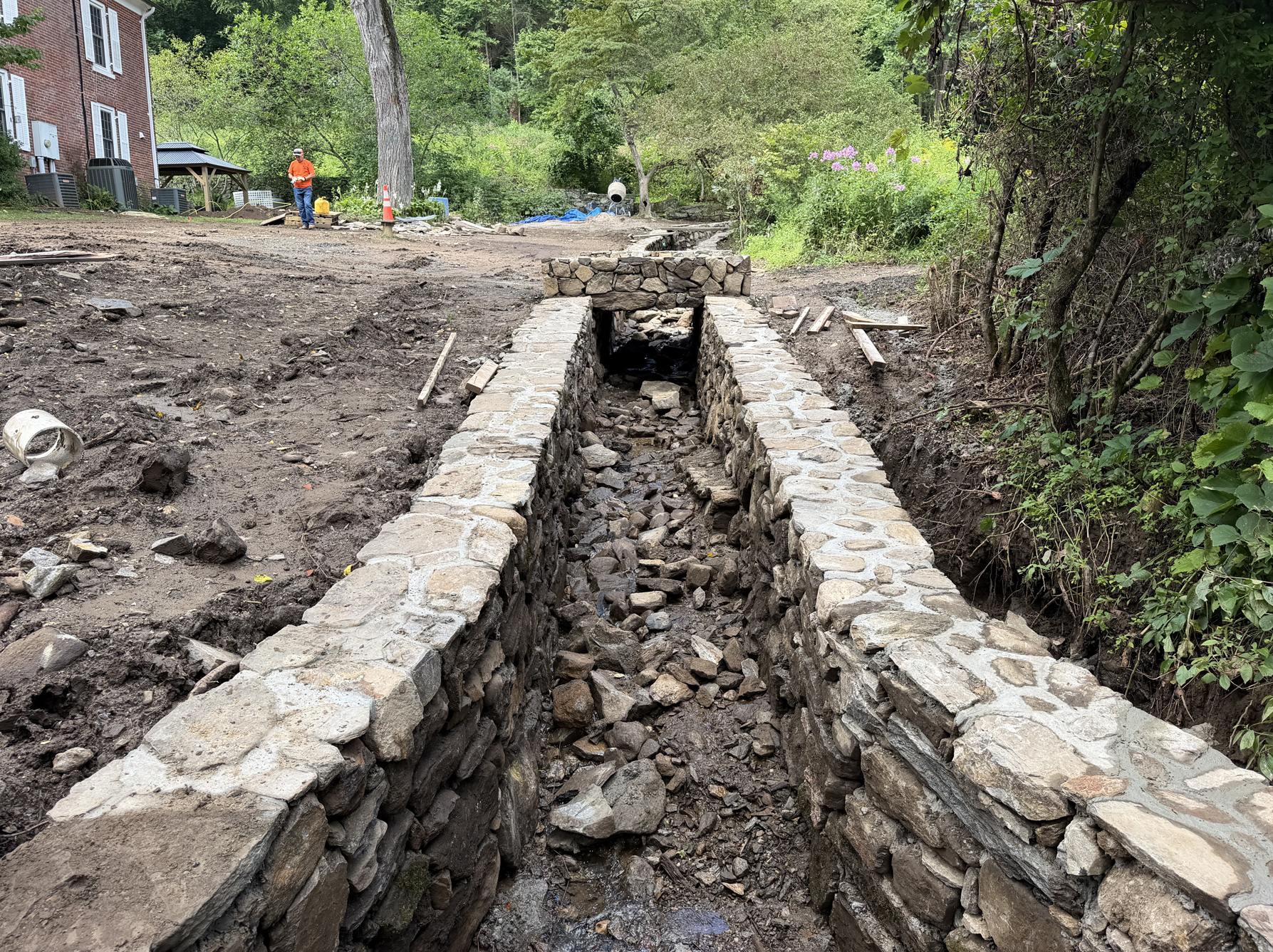 Stone-lined drainage ditch under construction in a muddy, wooded area