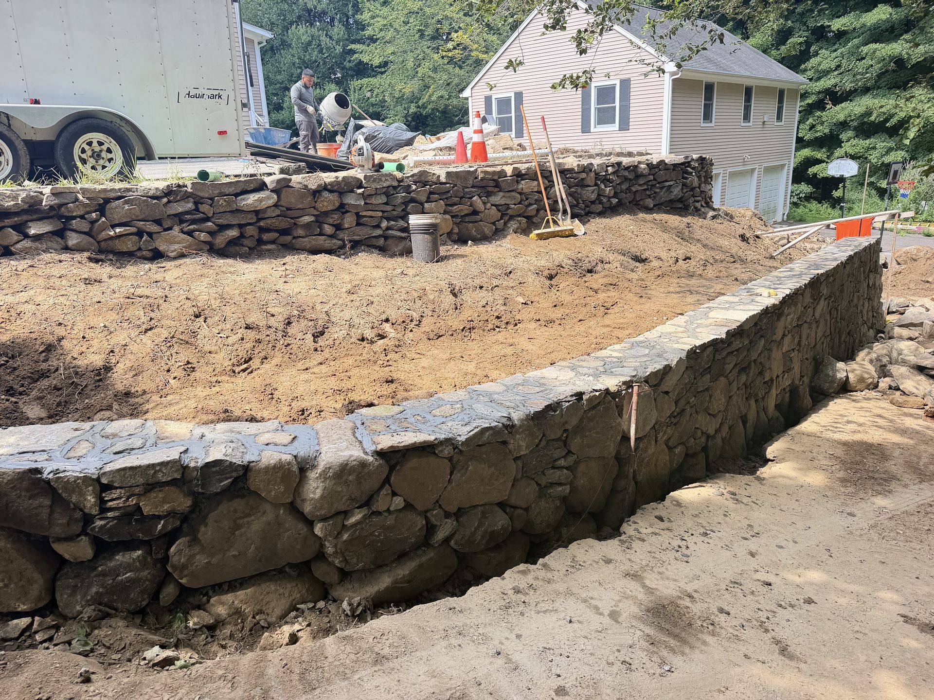 Curved stone retaining wall around a dirt lot at a construction site, with a house and truck in the background