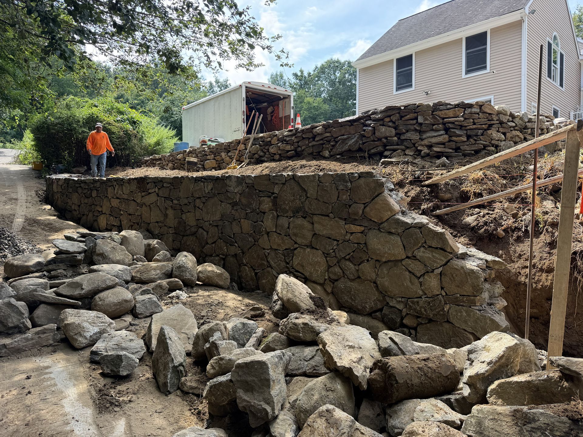 Stone retaining wall under construction beside a house, with worker in orange on a rocky slope.