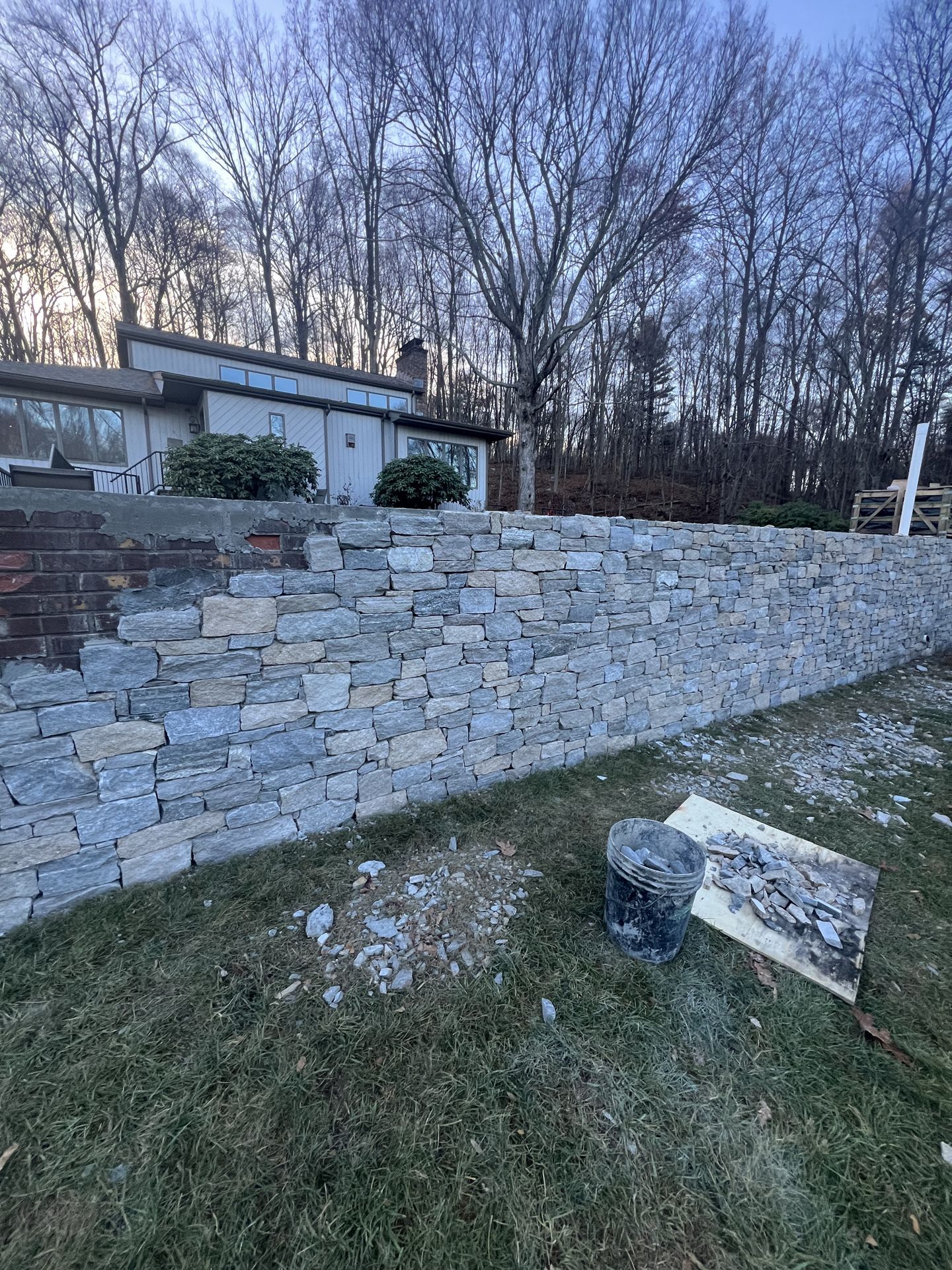 Stone retaining wall beside grassy yard and house at dusk, with trees in the background