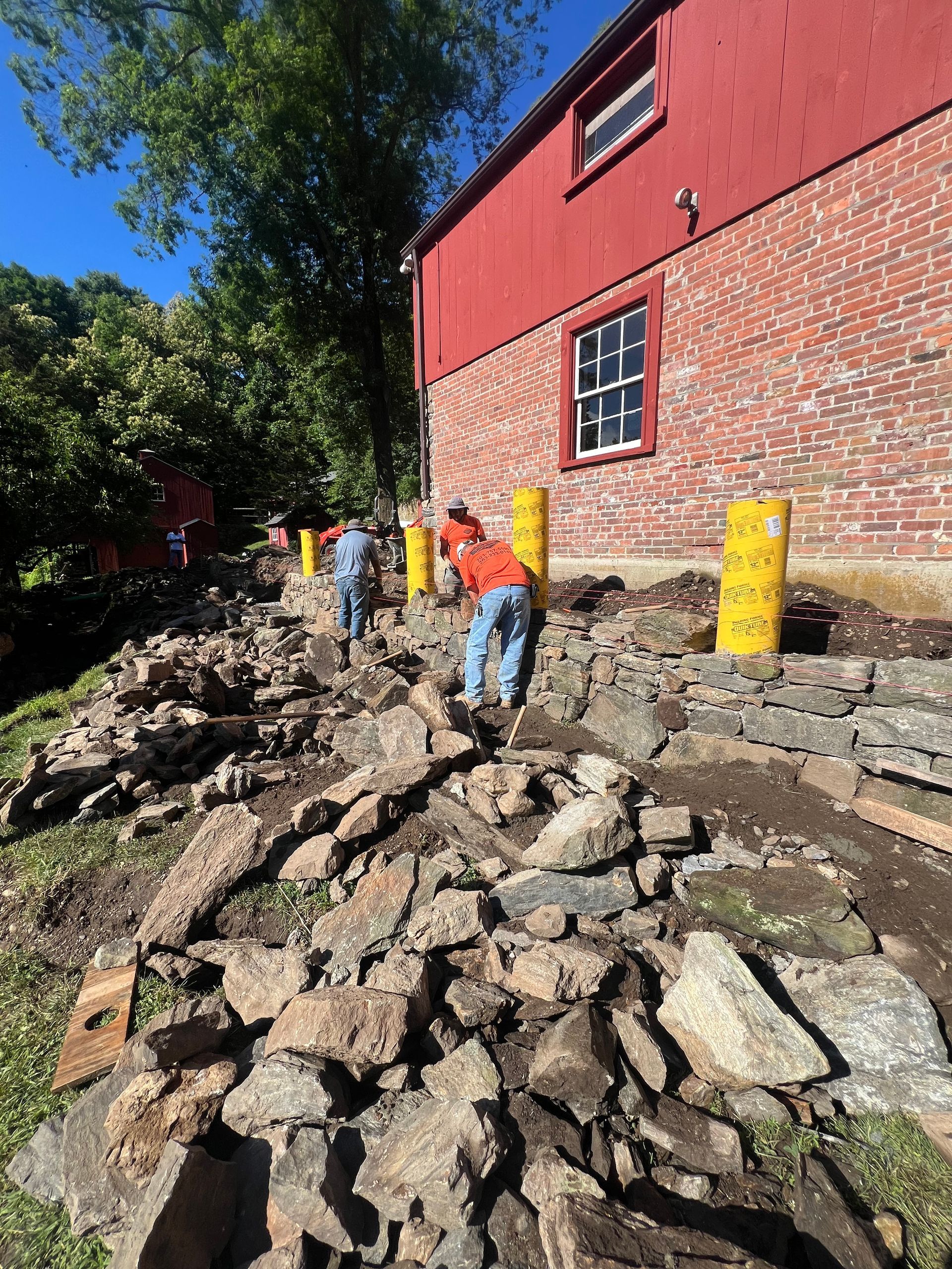 Workers in orange vests on a rocky path beside a red brick building and yellow posts