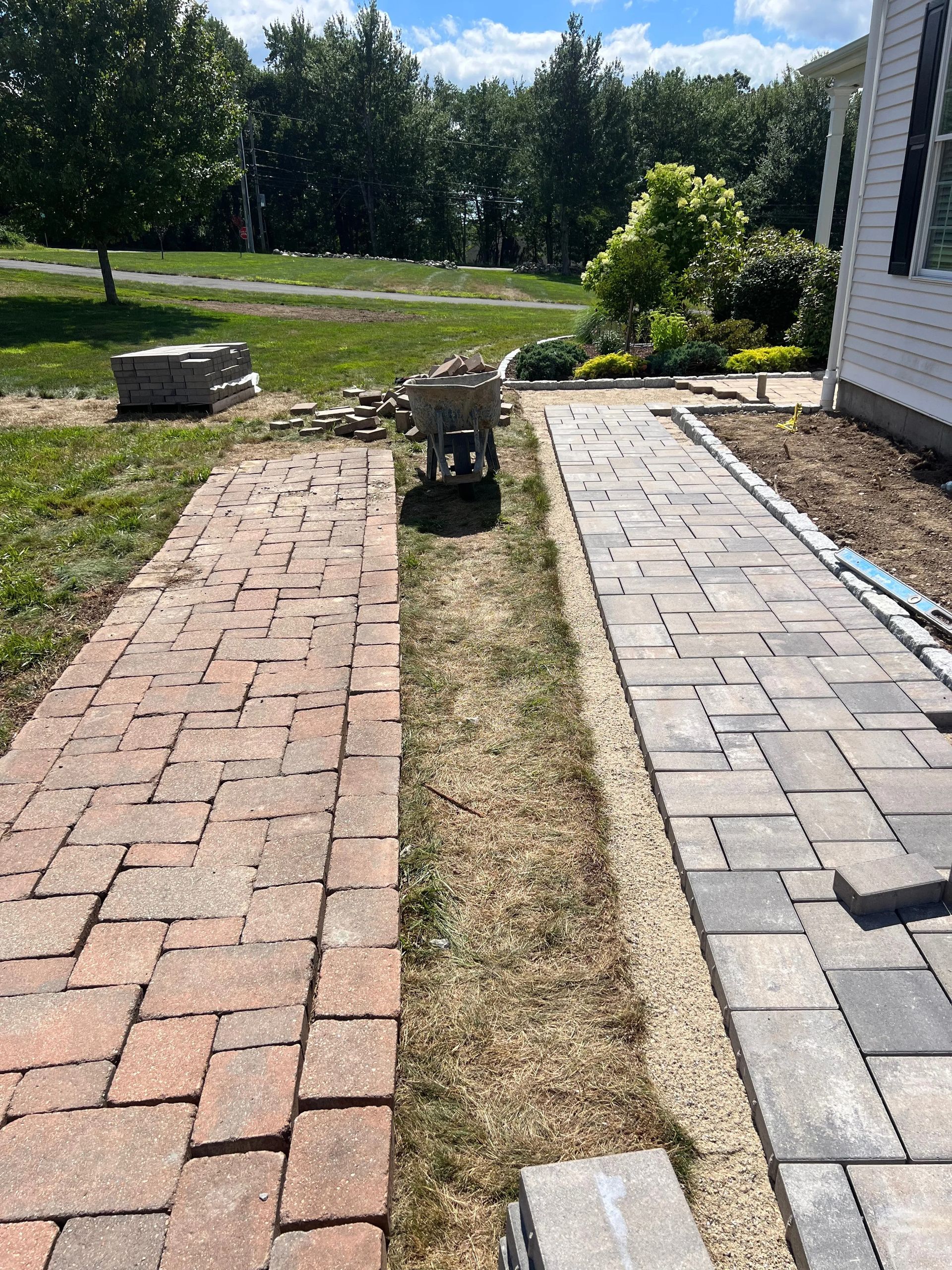 Brick walkway and paver path with a narrow dirt strip between them in a sunny yard.