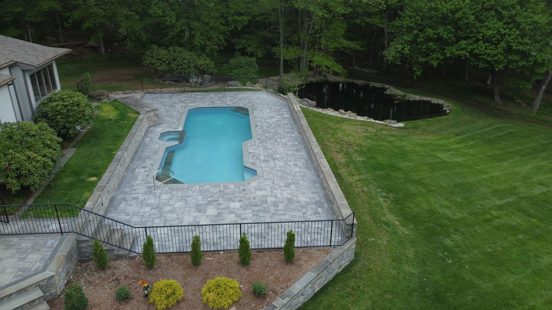 Aerial view of a backyard pool surrounded by stone patio and green lawn, bordered by trees and shrubs