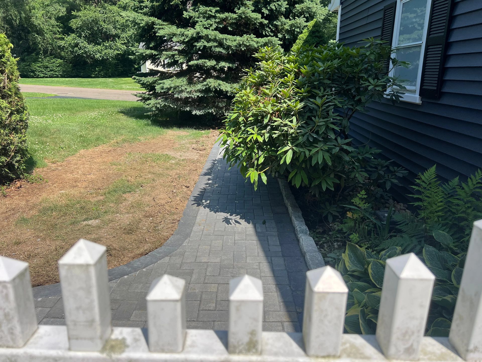 White picket fence overlooking a gray paver walkway beside a house and green lawn, with shrubs and trees nearby