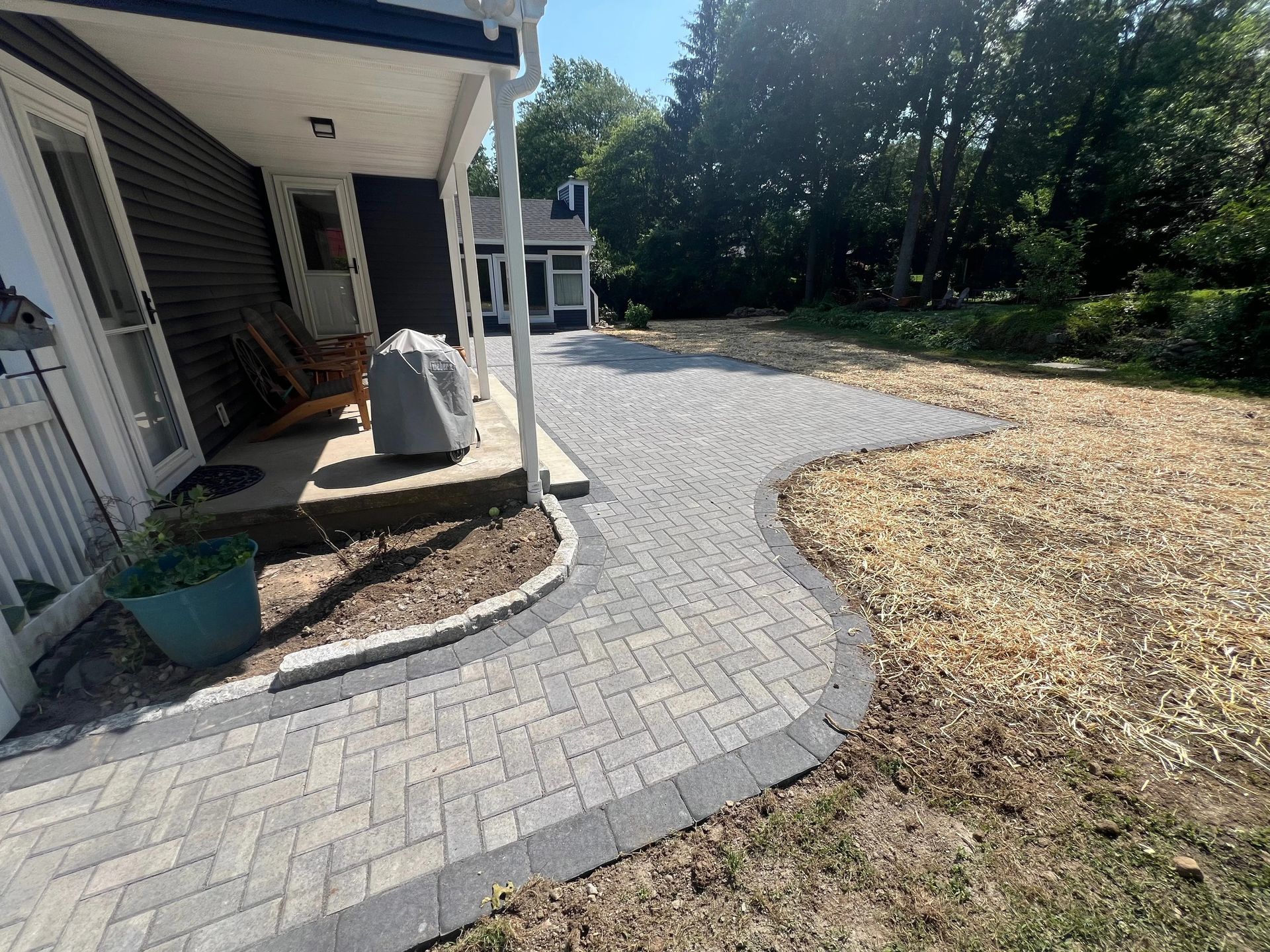Curved gray paver walkway beside a house, leading to a gravel driveway and trees beyond.